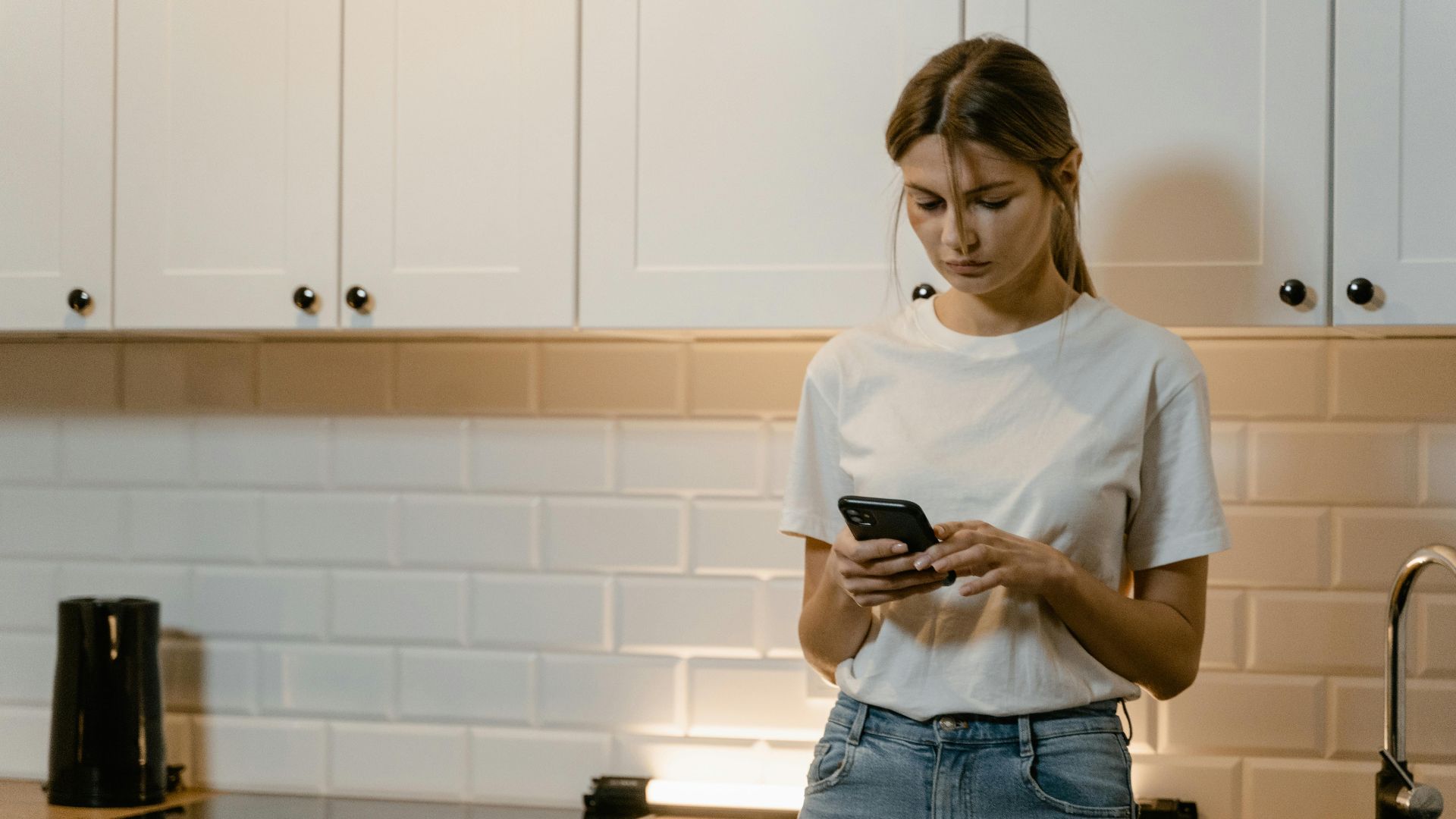 Young woman in kitchen using smartphone, looking focused and thoughtful.
