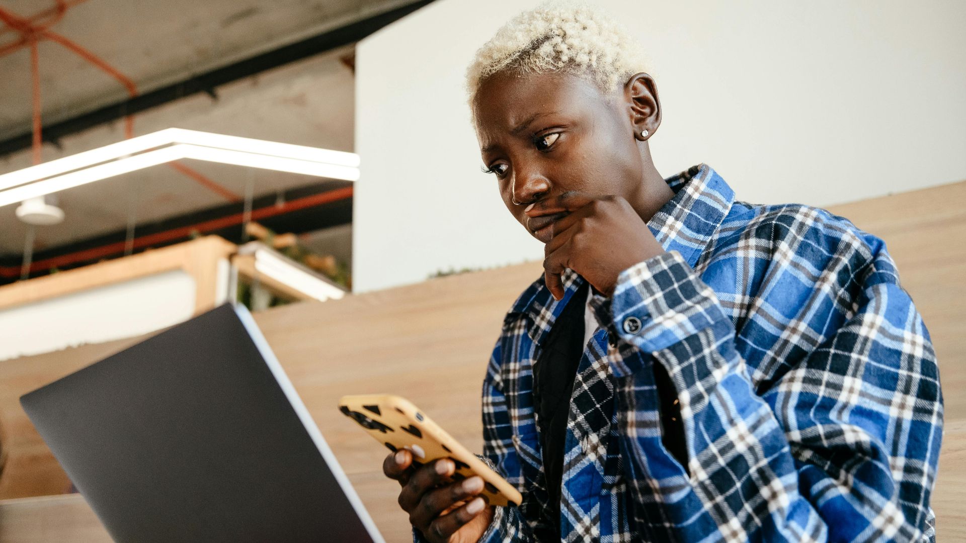 Young serious African American female in blue checkered shirt looking at screen of netbook while texting message on mobile phone