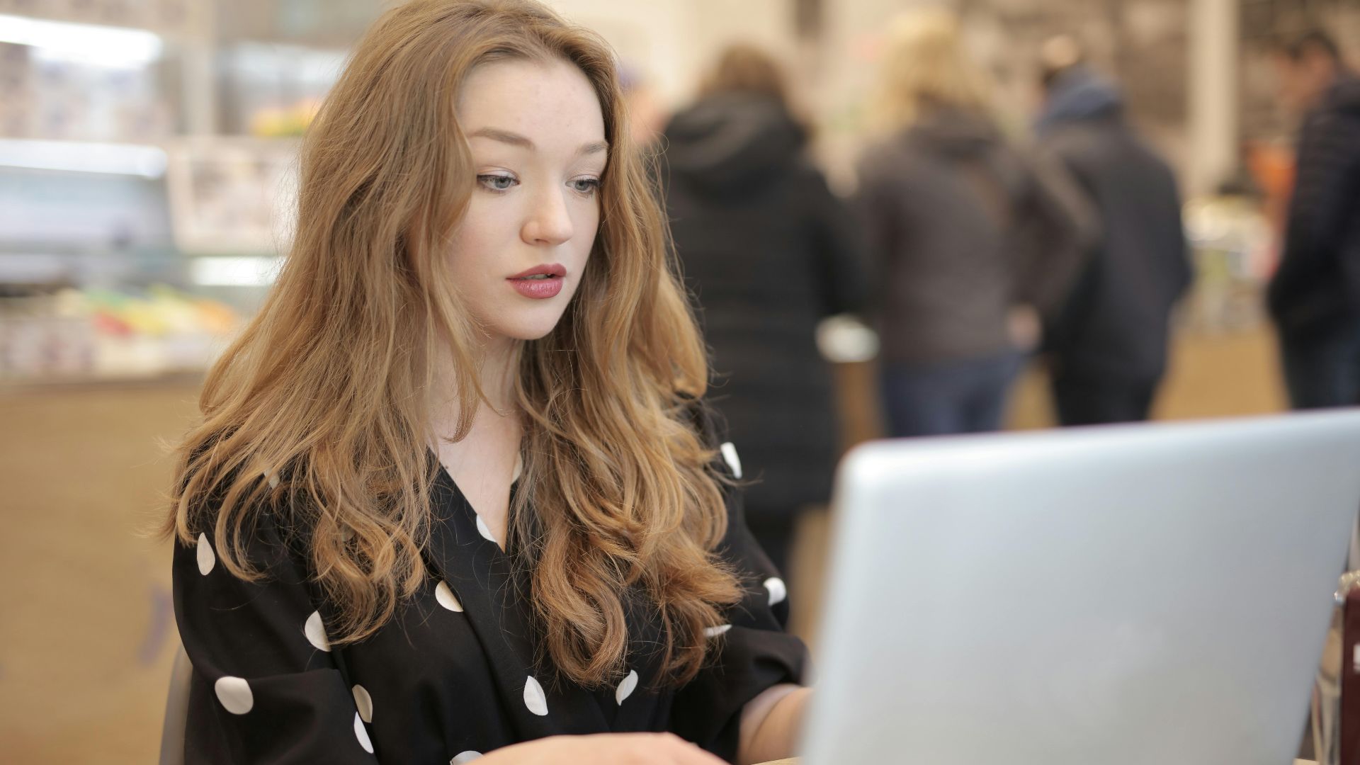 A focused woman in a polka dot shirt is typing on a laptop in a bustling cafe.