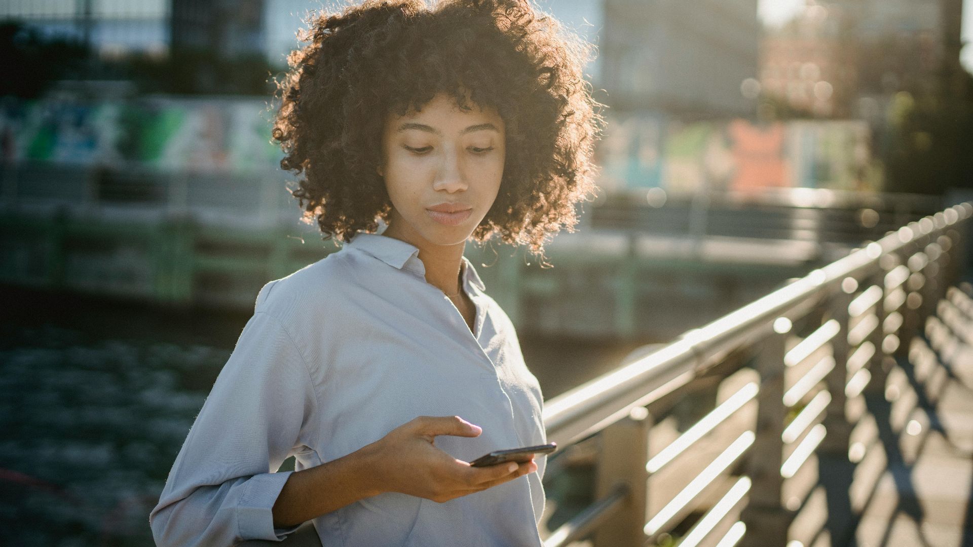 Portrait of a young woman with curly hair using her smartphone on an urban bridge during daylight.