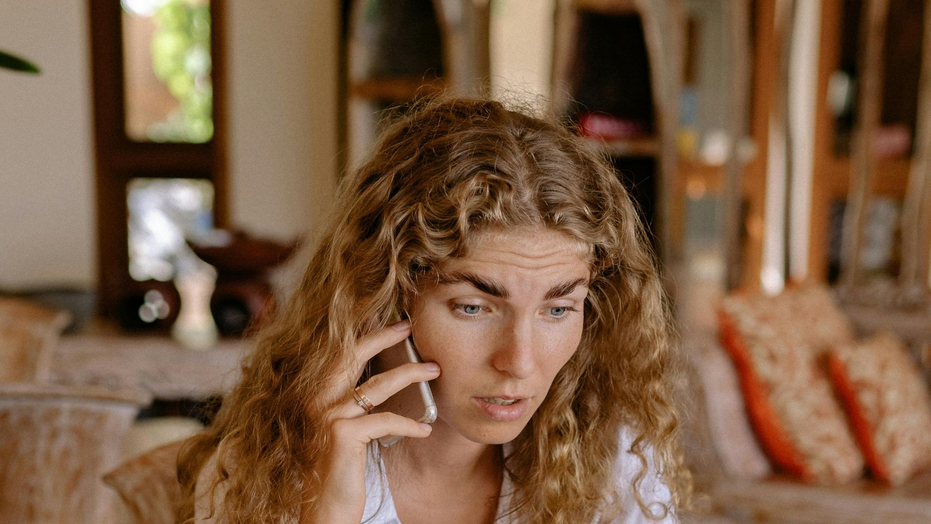 Woman sitting at wooden table, using laptop and talking on phone in a cozy indoor setting.