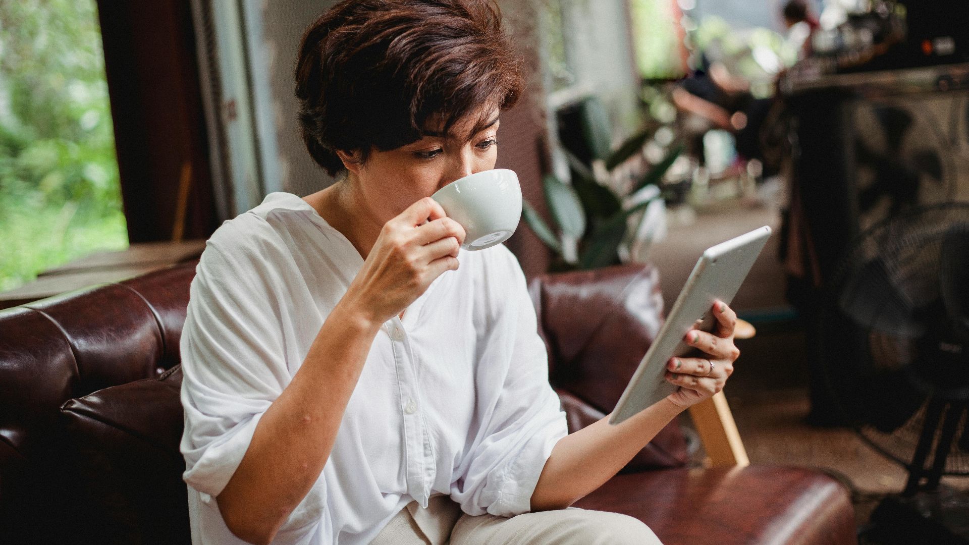 Focused Asian female in trendy clothes drinking cup of beverage and looking at screen of tablet