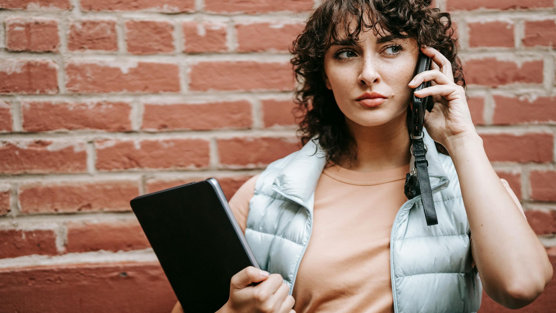 Adult attentive female remote worker with netbook talking on cellphone while looking away against brick wall