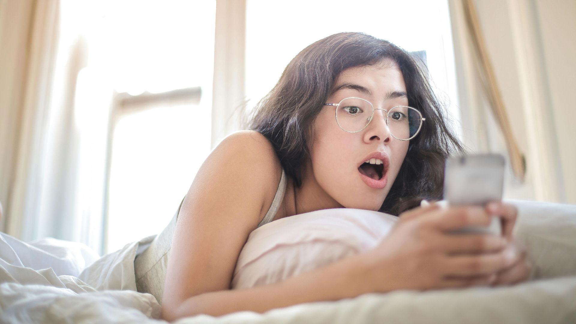 Young woman lying on bed, surprised by something on her smartphone, in a bright bedroom setting.