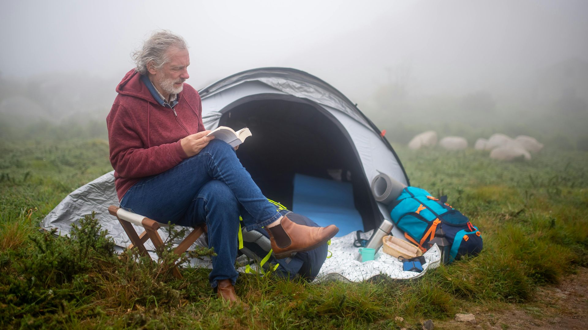 A senior man enjoys reading at a campsite in Portugal's lush landscape, surrounded by mist.