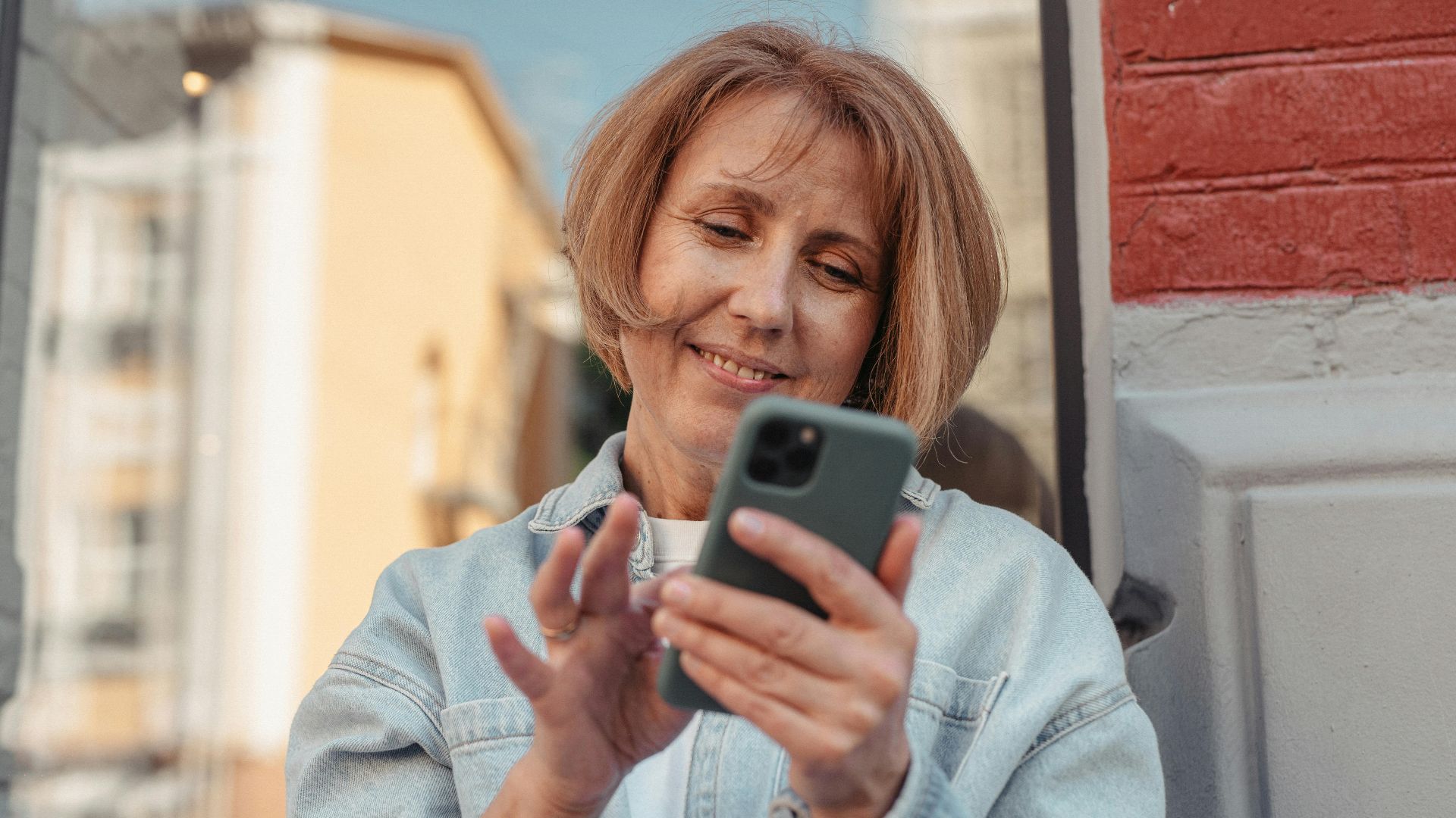 A middle-aged woman in a denim jacket smiles while using her smartphone outside.