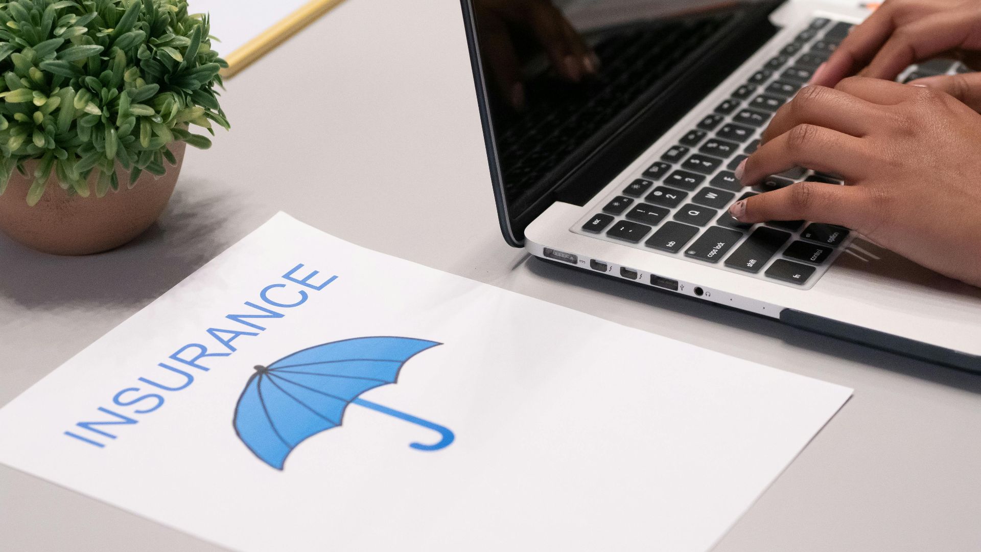 Close-up of hands typing on laptop with an insurance document visible on the desk.