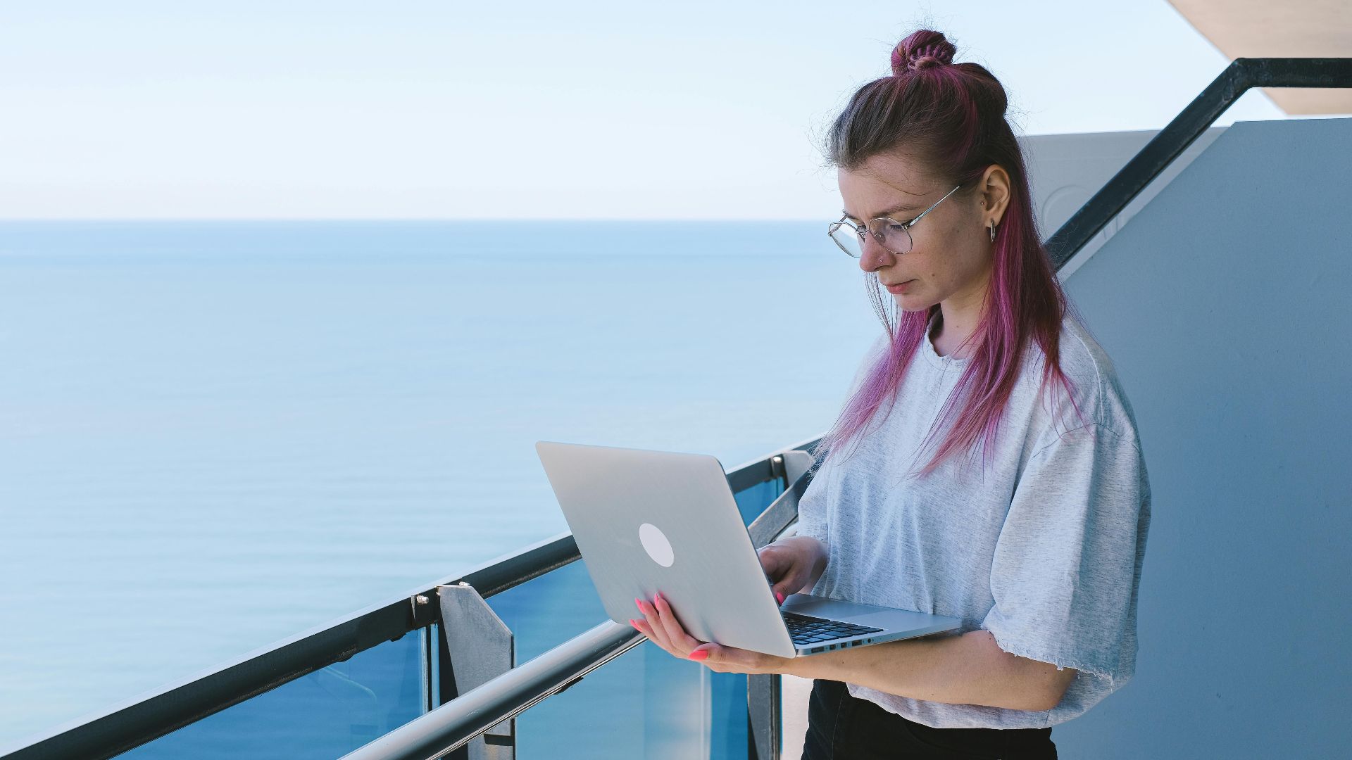 A young woman works remotely on her laptop by the sea on a sunny day, embodying the digital nomad lifestyle.