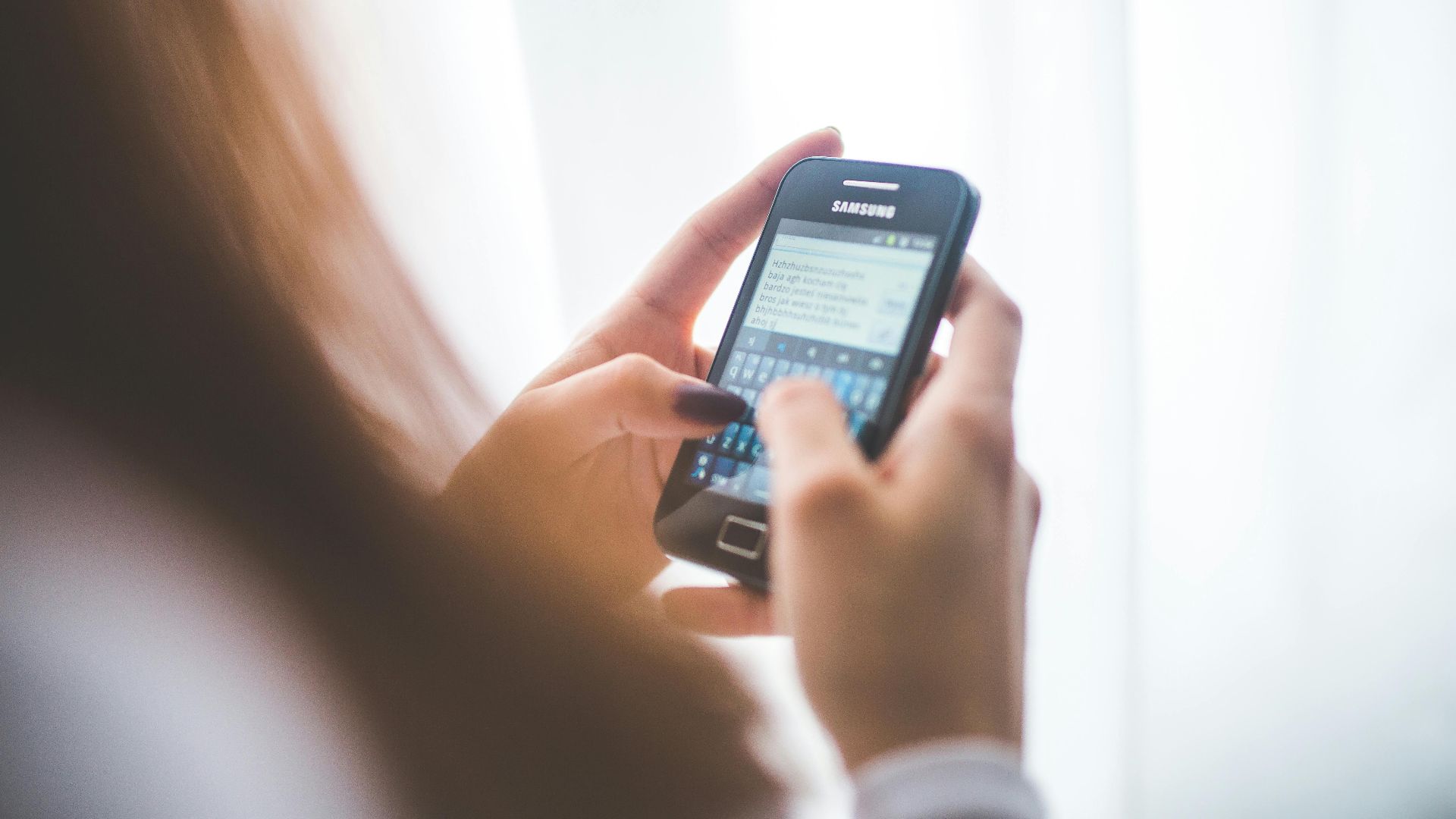 Close-up of a young woman typing on a Samsung smartphone at home. Bright indoor lighting.