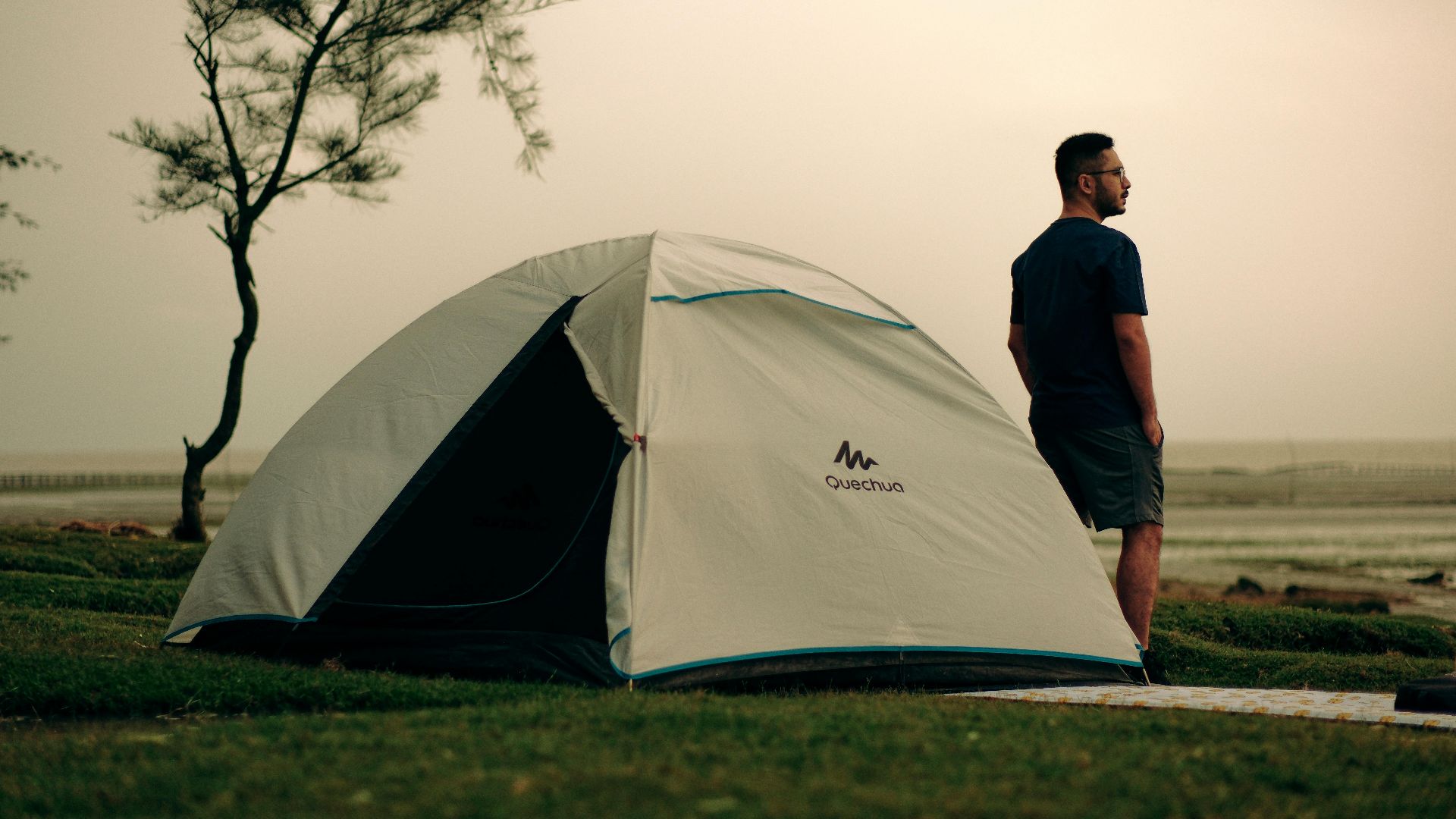 A man standing by a tent on a coastal campsite, capturing a serene sunset moment.