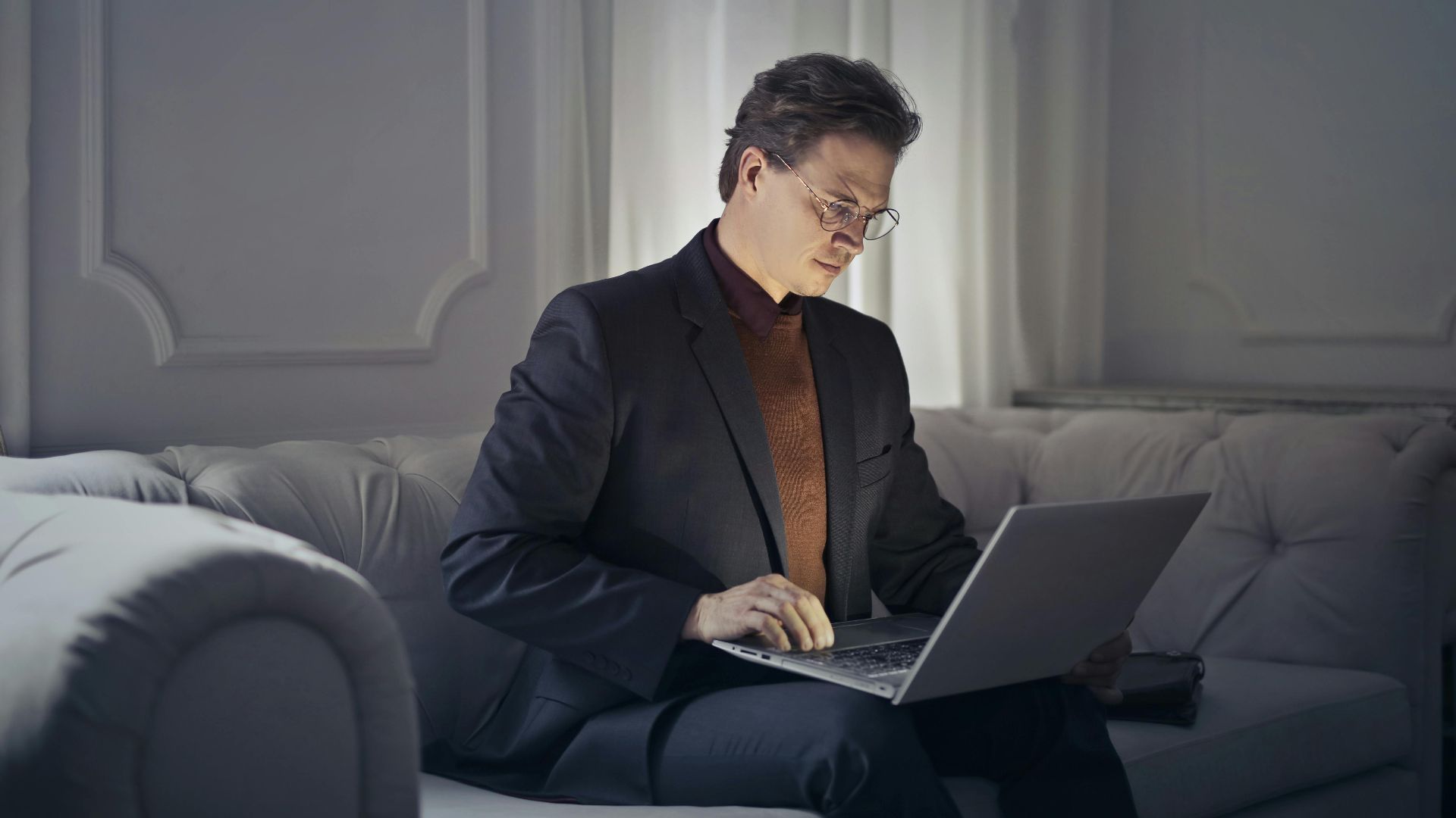 Professional man sitting on sofa using laptop for remote work in a stylish indoor setting.