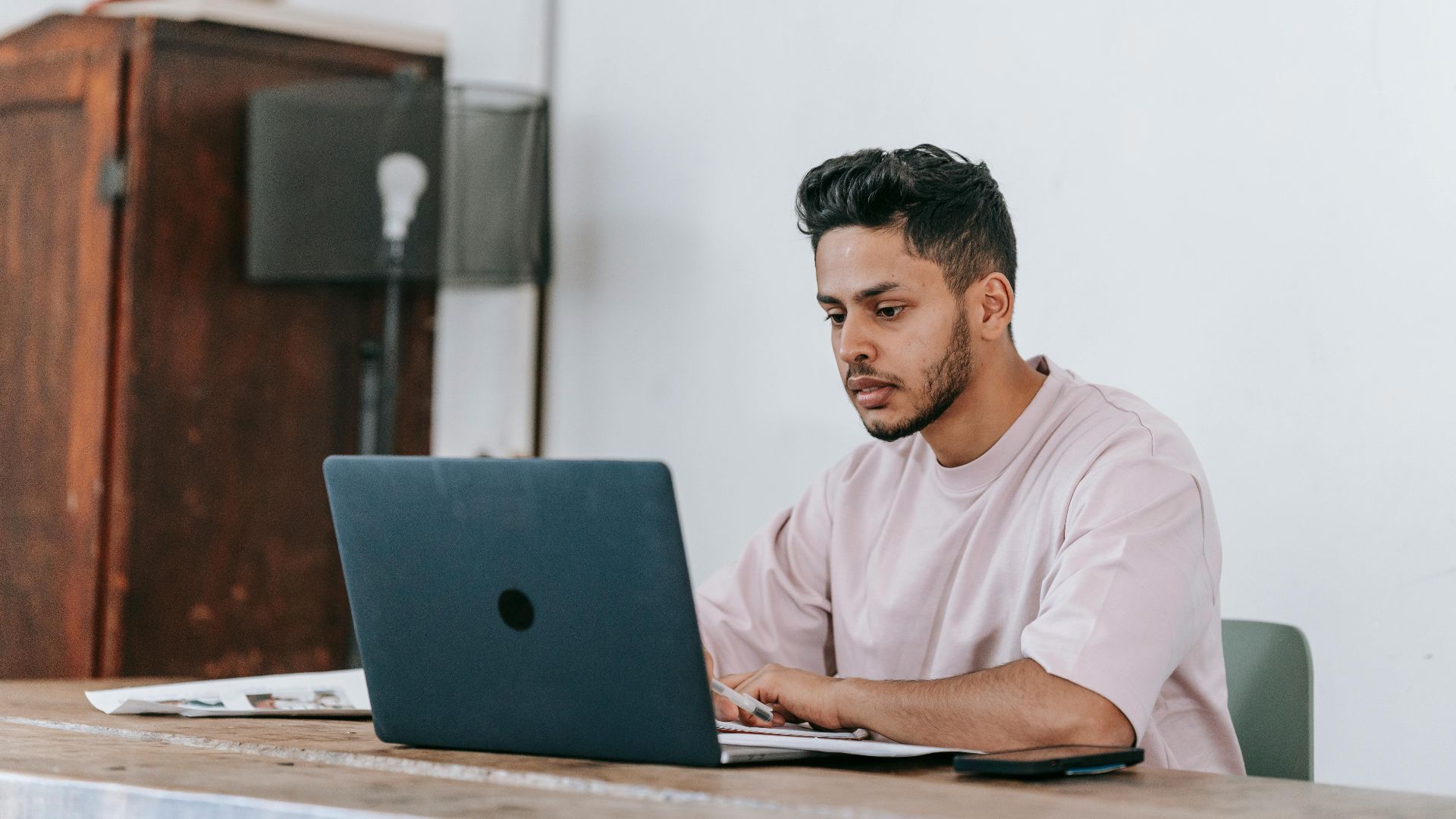 Concentrated young ethnic male distance employee with modern haircut working on netbook while sitting at desk