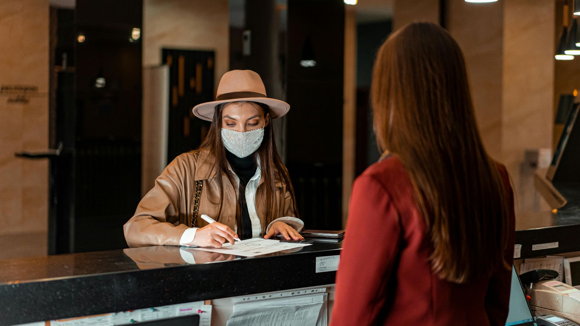 A woman in a hat and face mask checks in at a hotel reception, interacting with a receptionist.