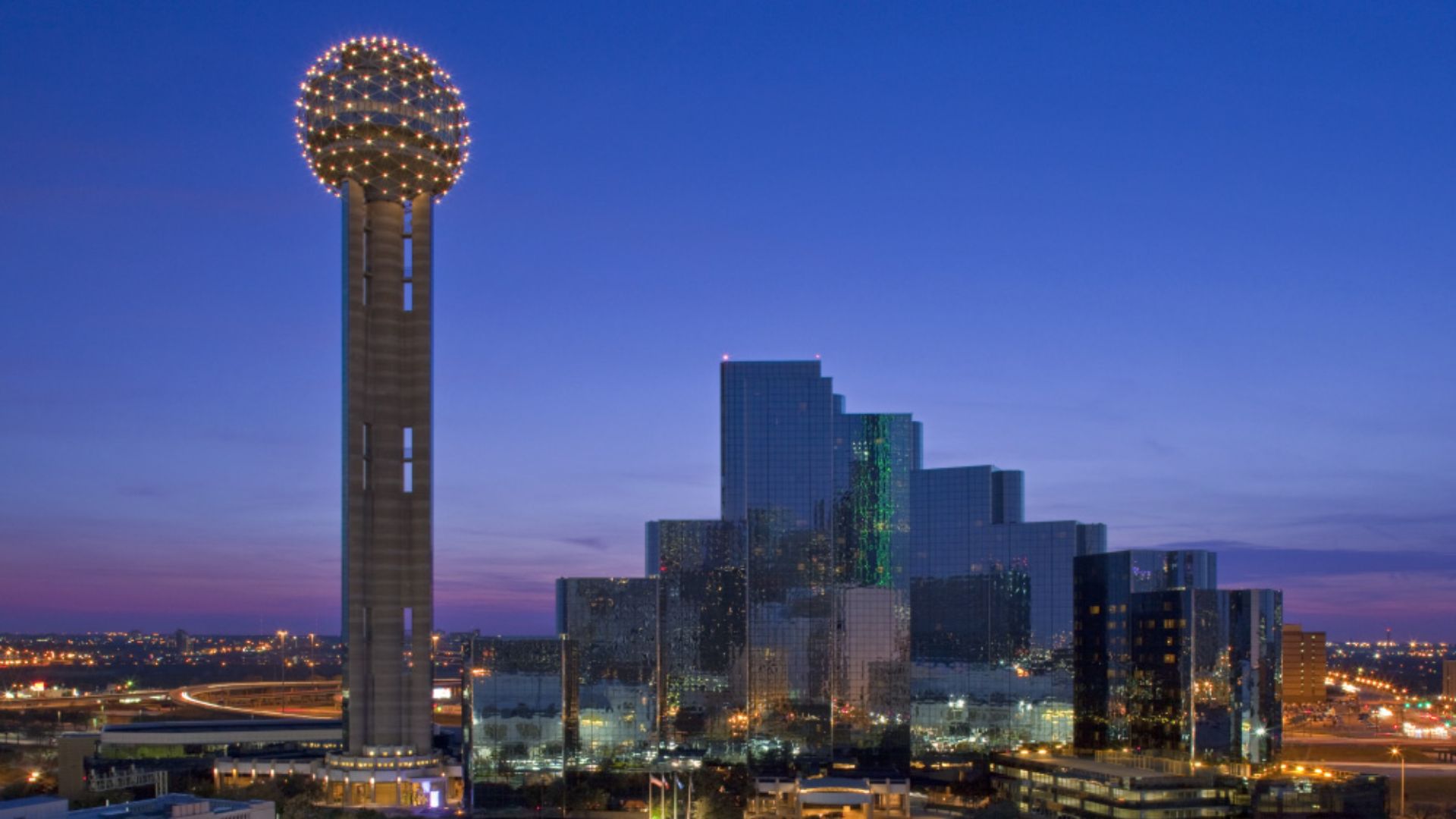 This image shows the iconic skyline of Downtown Dallas. You can clearly see the Reunion Tower which is connected to Hyatt Regency Dallas. The lights in the tower are aglow during this sunset shot taken from just outside of downtown.