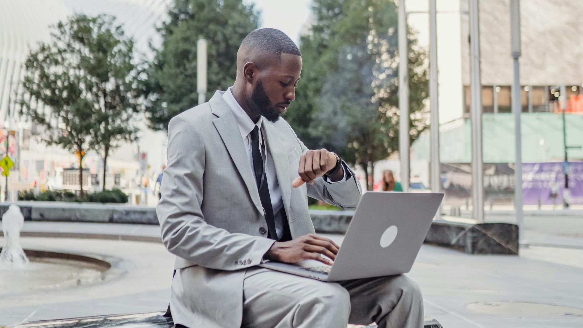A businessman in a suit checks his wristwatch while using a laptop outdoors in an urban setting.