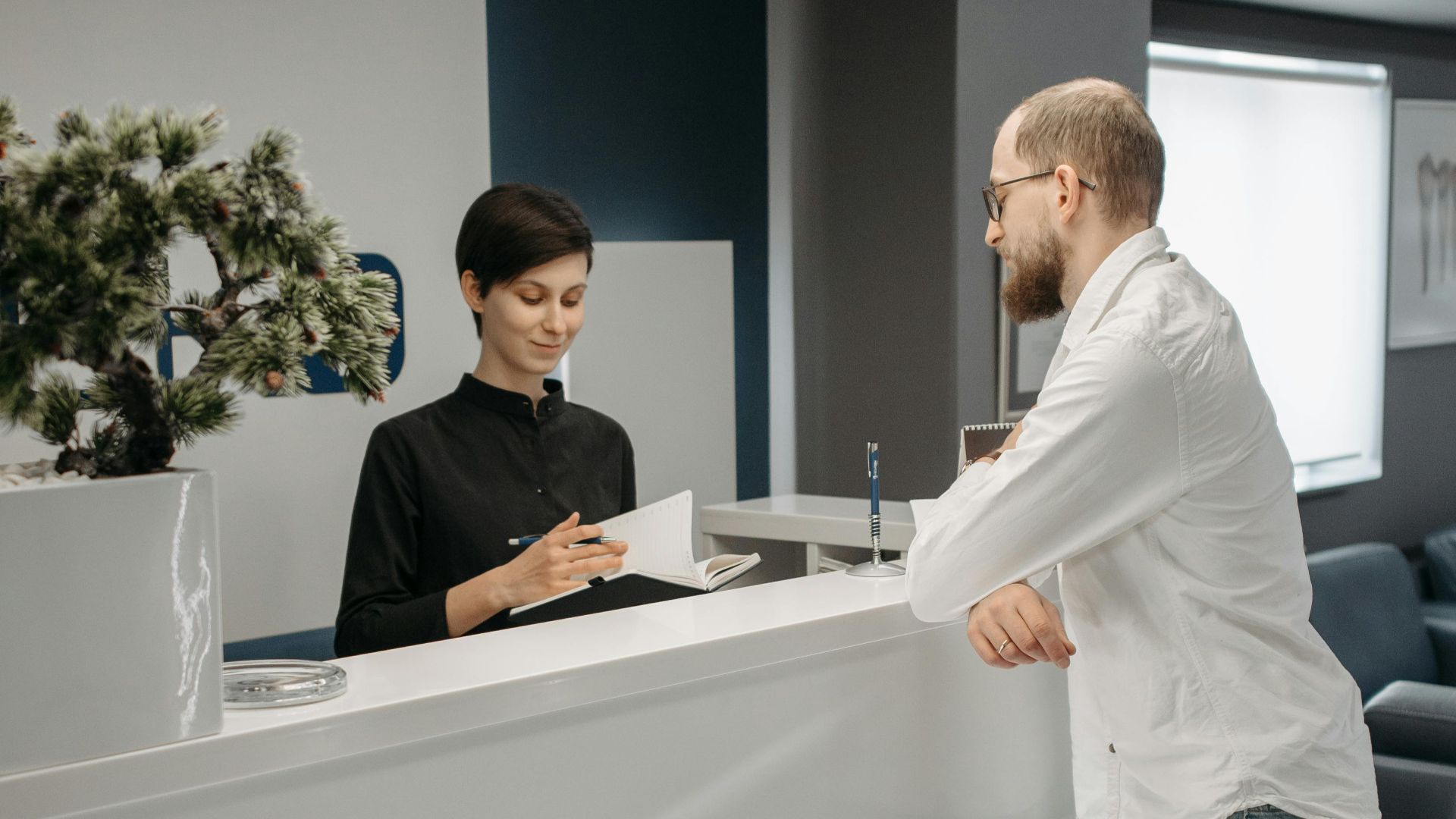 A man and woman at a sleek office reception desk, discussing paperwork in a professional setting.