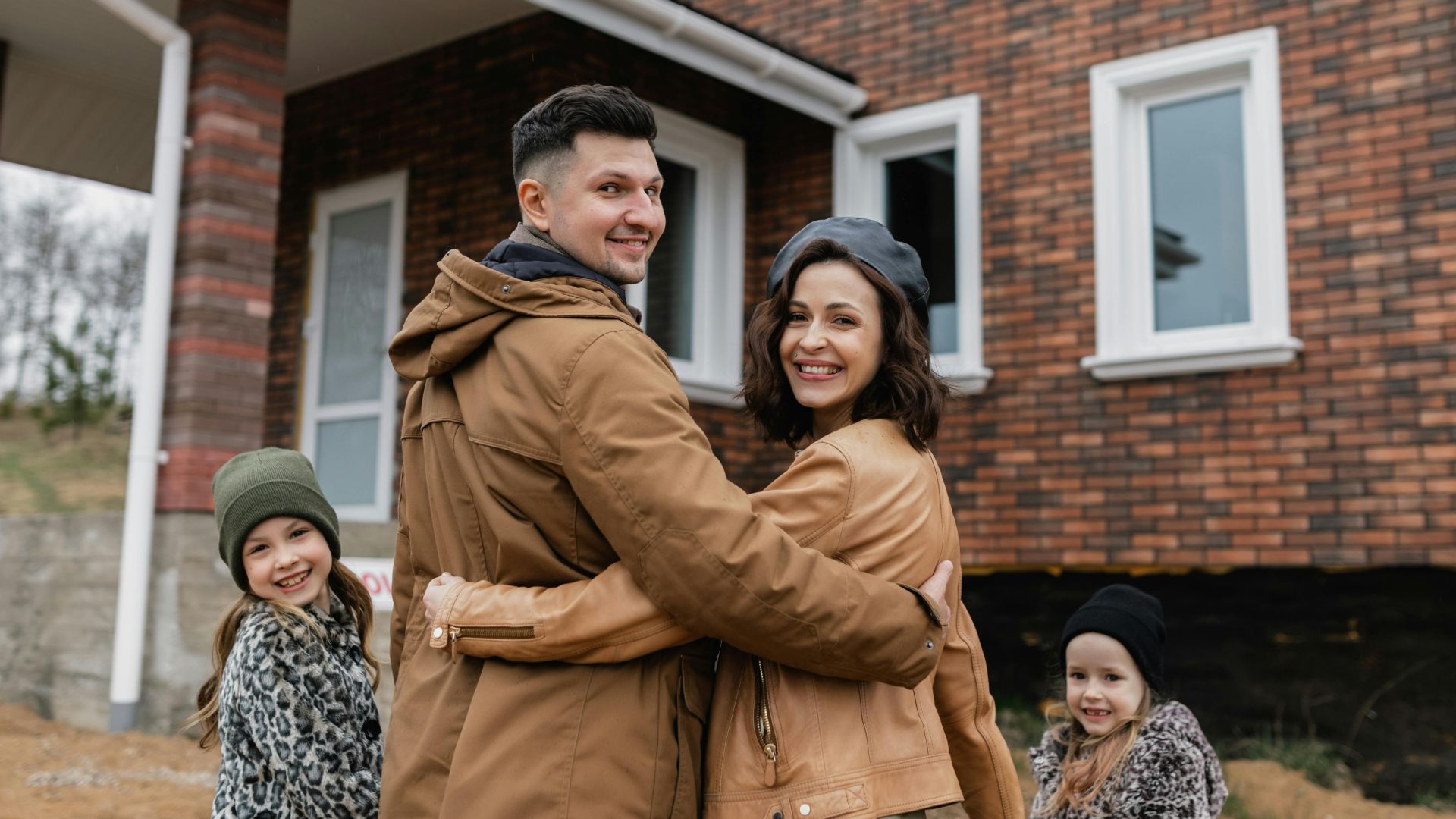 Family of four happily posing in front of their new brick house on a sunny day.