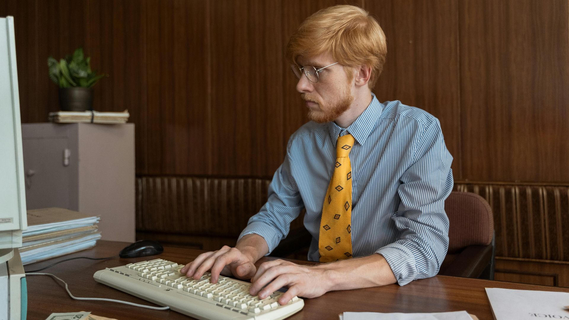 A man with red hair and eyeglasses typing on a retro keyboard in a vintage office setting.