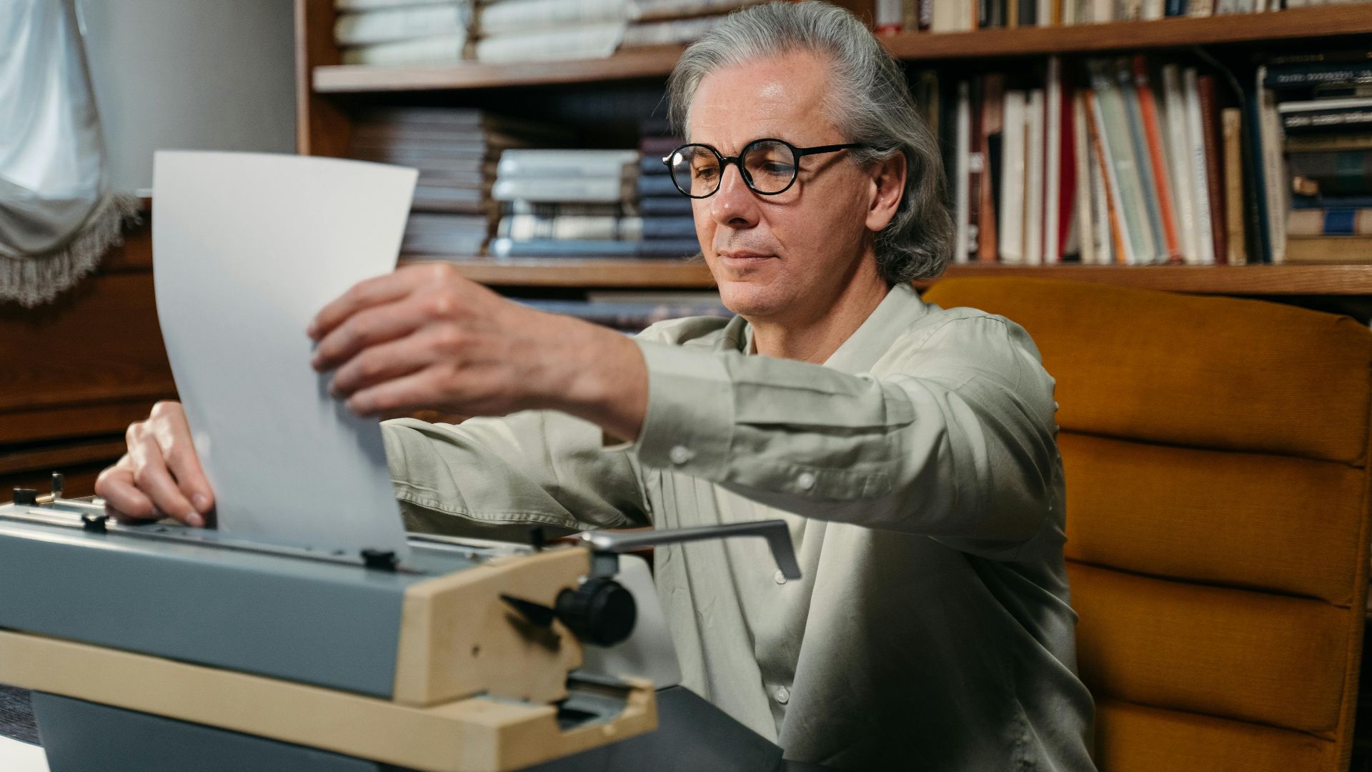 Senior man with glasses types on vintage typewriter in a library with bookshelves.