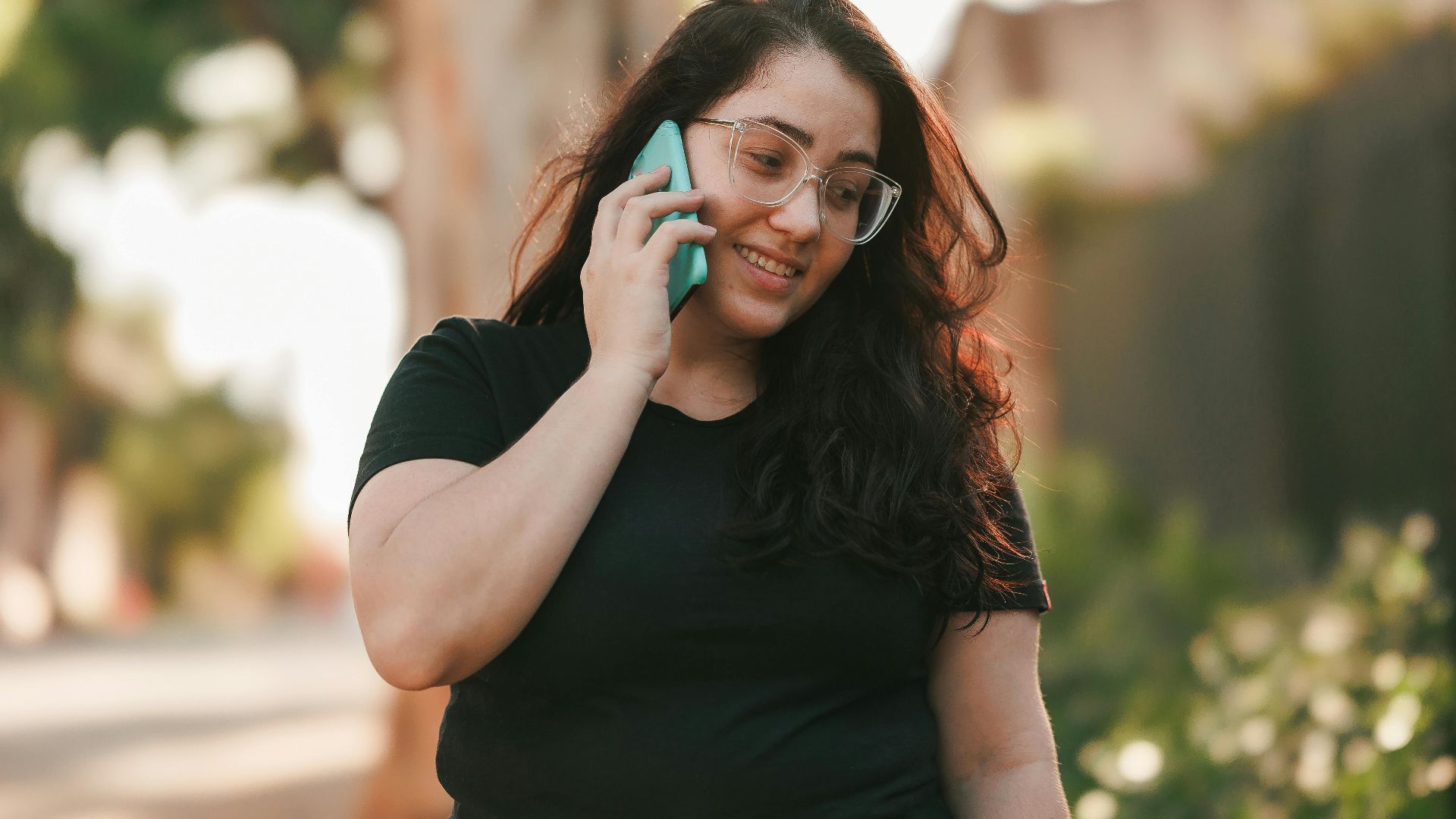 Smiling woman with glasses talking on her smartphone outside, enjoying a warm day.