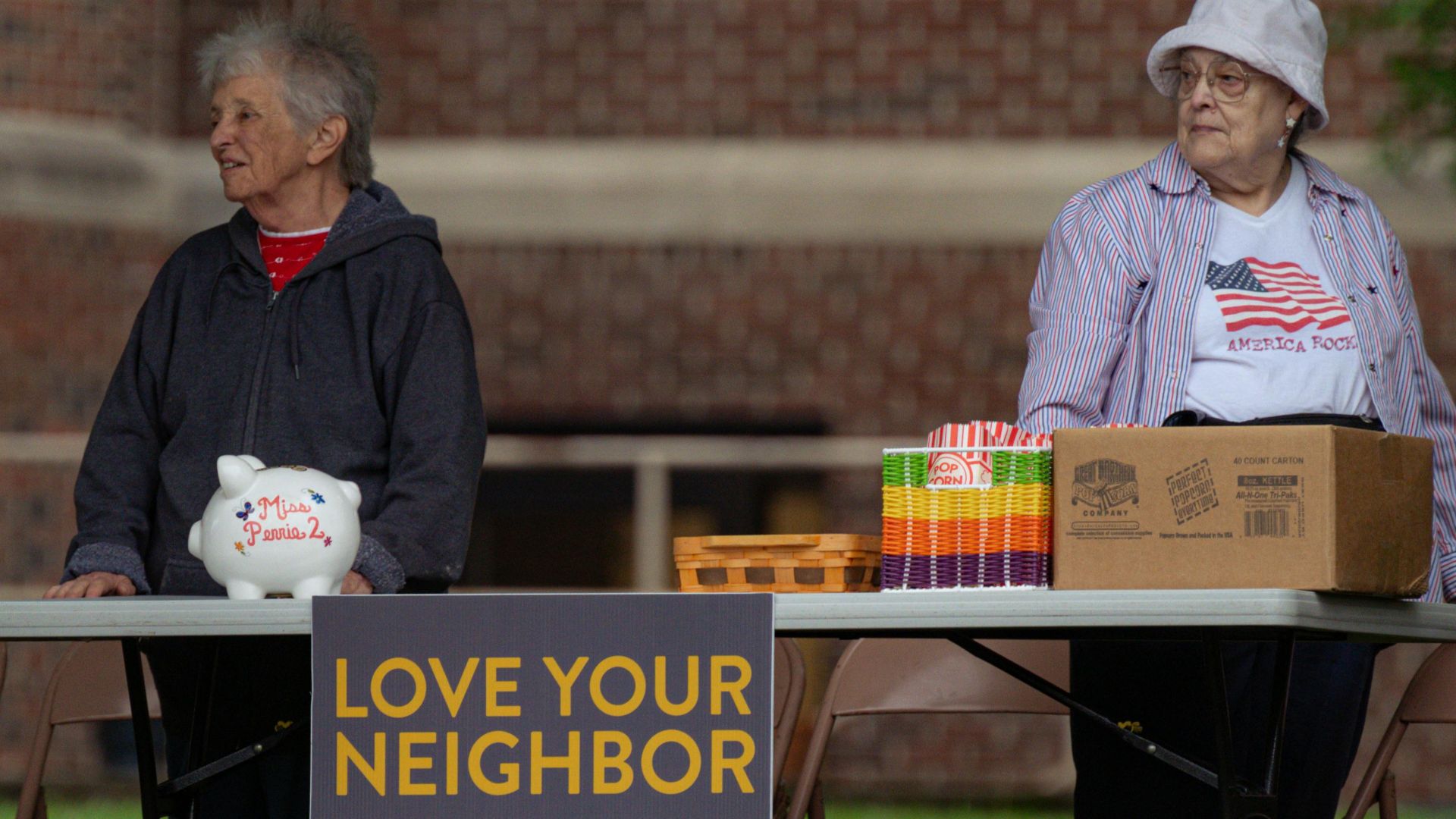 Two elderly women volunteer at a community event with a 'Love Your Neighbor' sign.