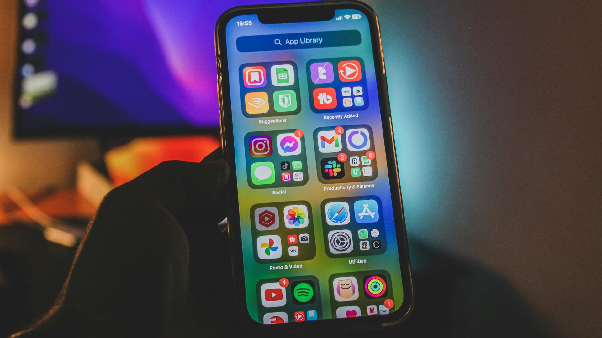 A hand holds a smartphone showing a colorful app-filled screen in an indoor room with dim lighting.