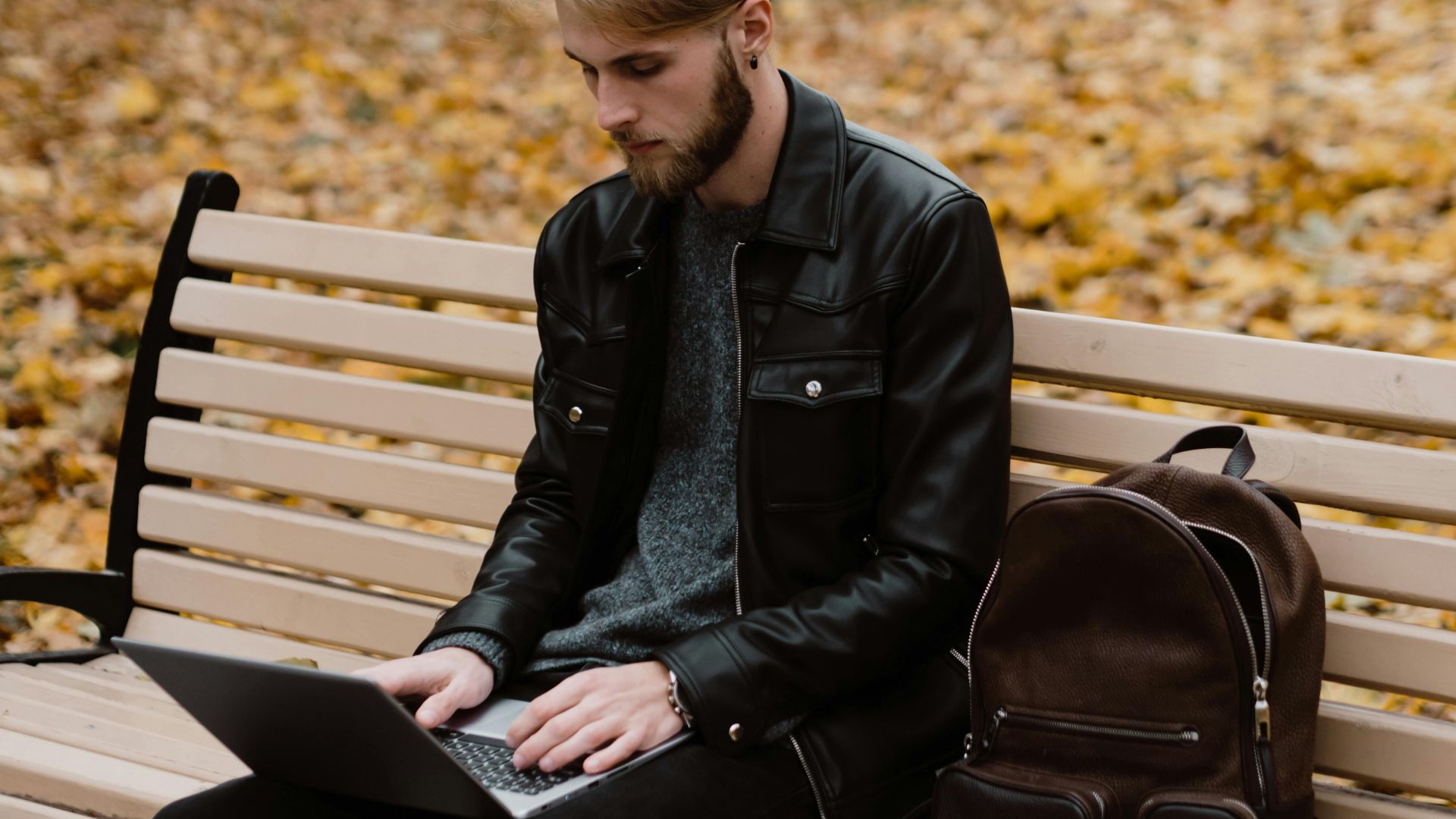 A man with manbun typing on a laptop while sitting on a park bench, surrounded by autumn leaves.