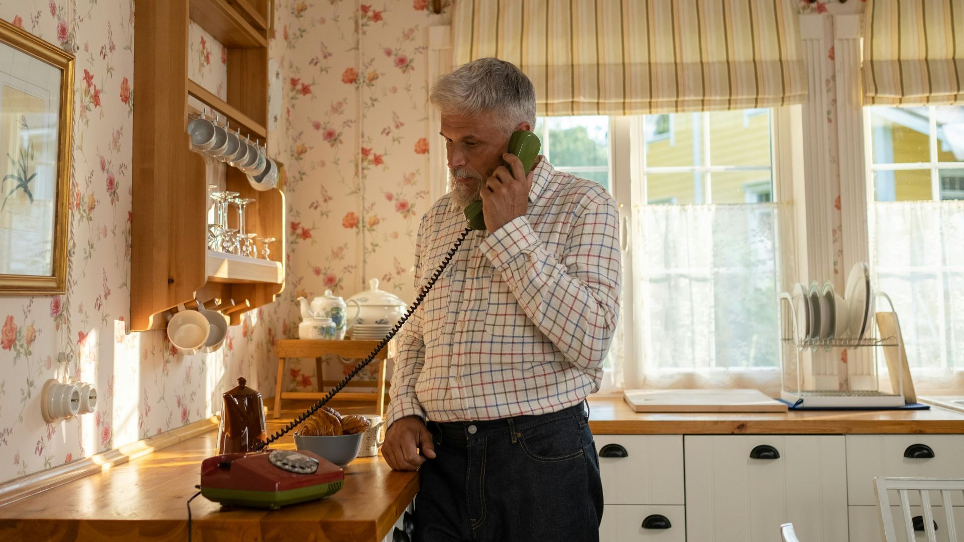 Senior man talks on a vintage rotary phone in a retro-styled kitchen with floral wallpaper and classic decor.