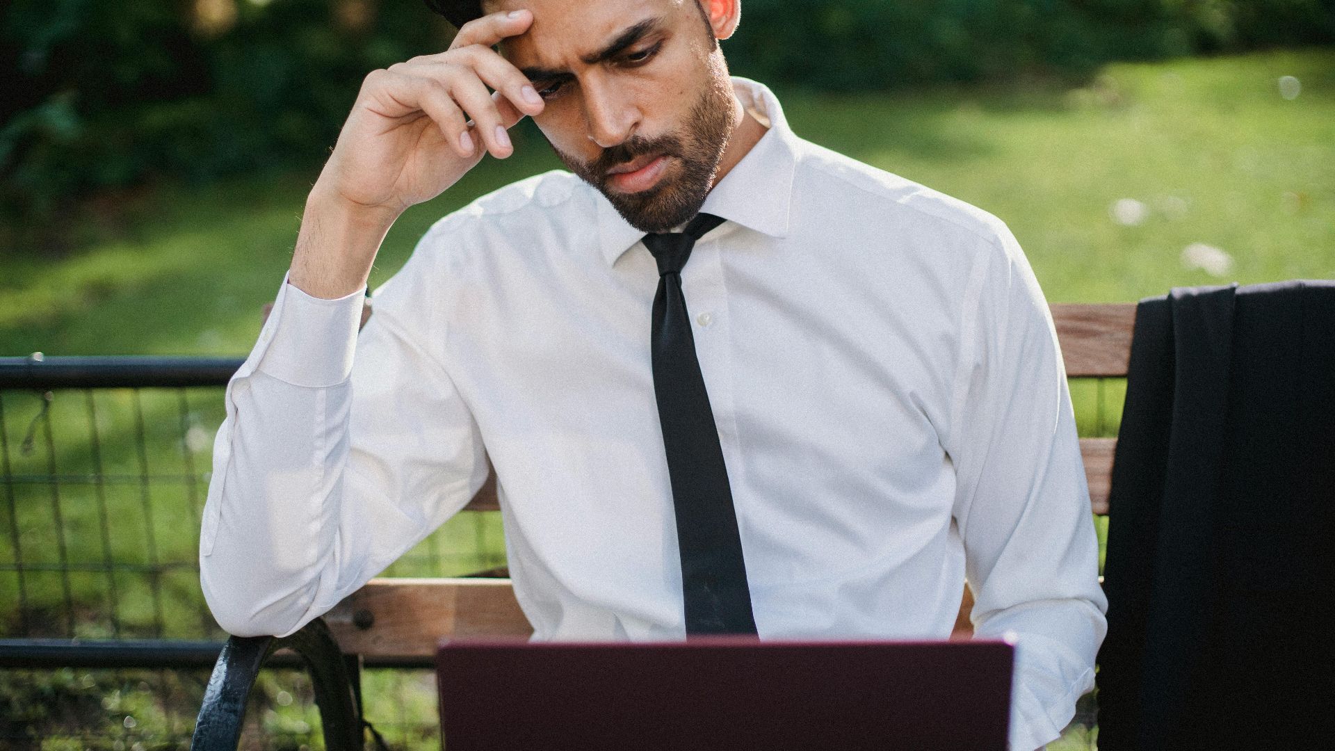 Bearded man in a white shirt and tie working on a laptop on a park bench.