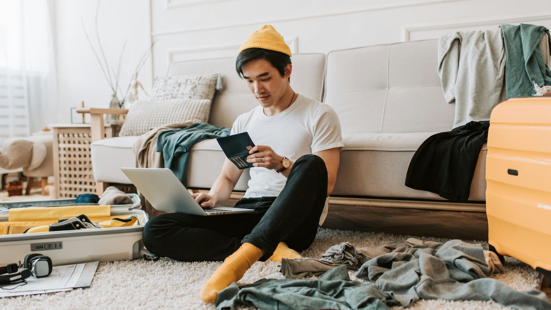 A young man in a messy living room preparing for travel with a laptop and passport.