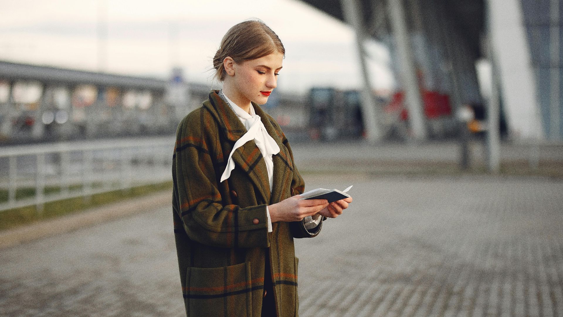Attentive female passenger wearing trendy plaid coat and white blouse checking passport and ticket standing on pavement near modern building of airport outside