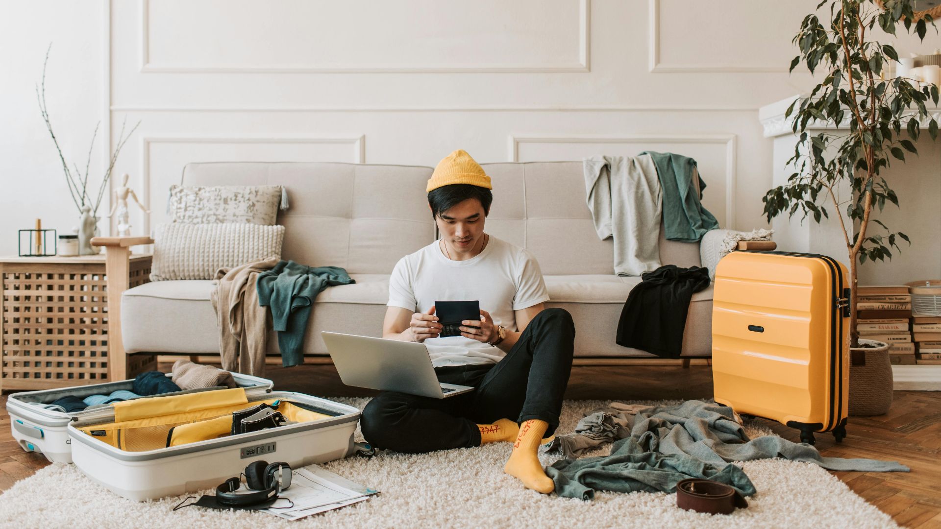 A young man uses multiple gadgets while sitting in a stylish, cozy living room with a yellow suitcase and open luggage.