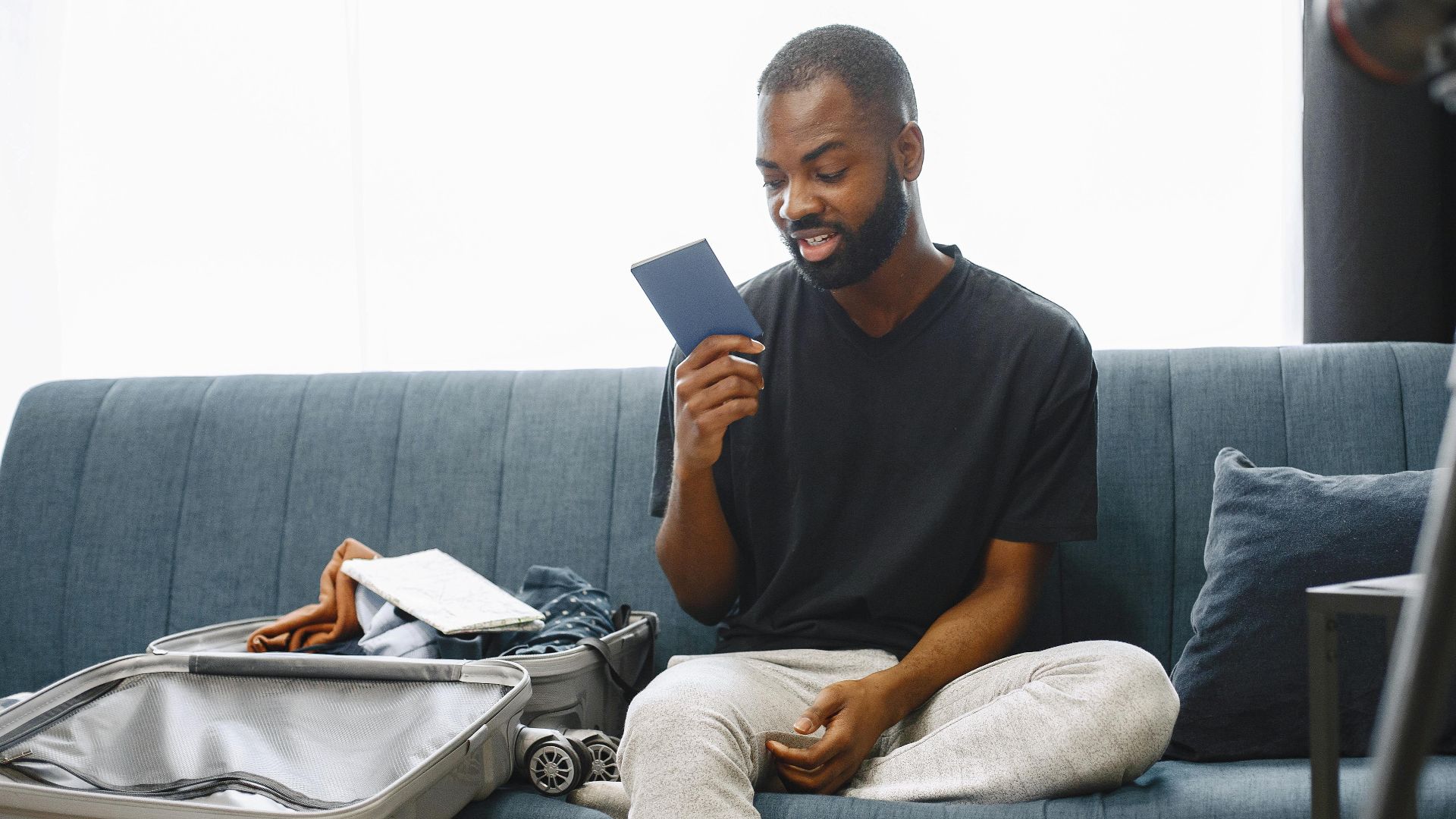 Adult man sits on sofa with open luggage, holding passport, preparing for travel, vlogging.
