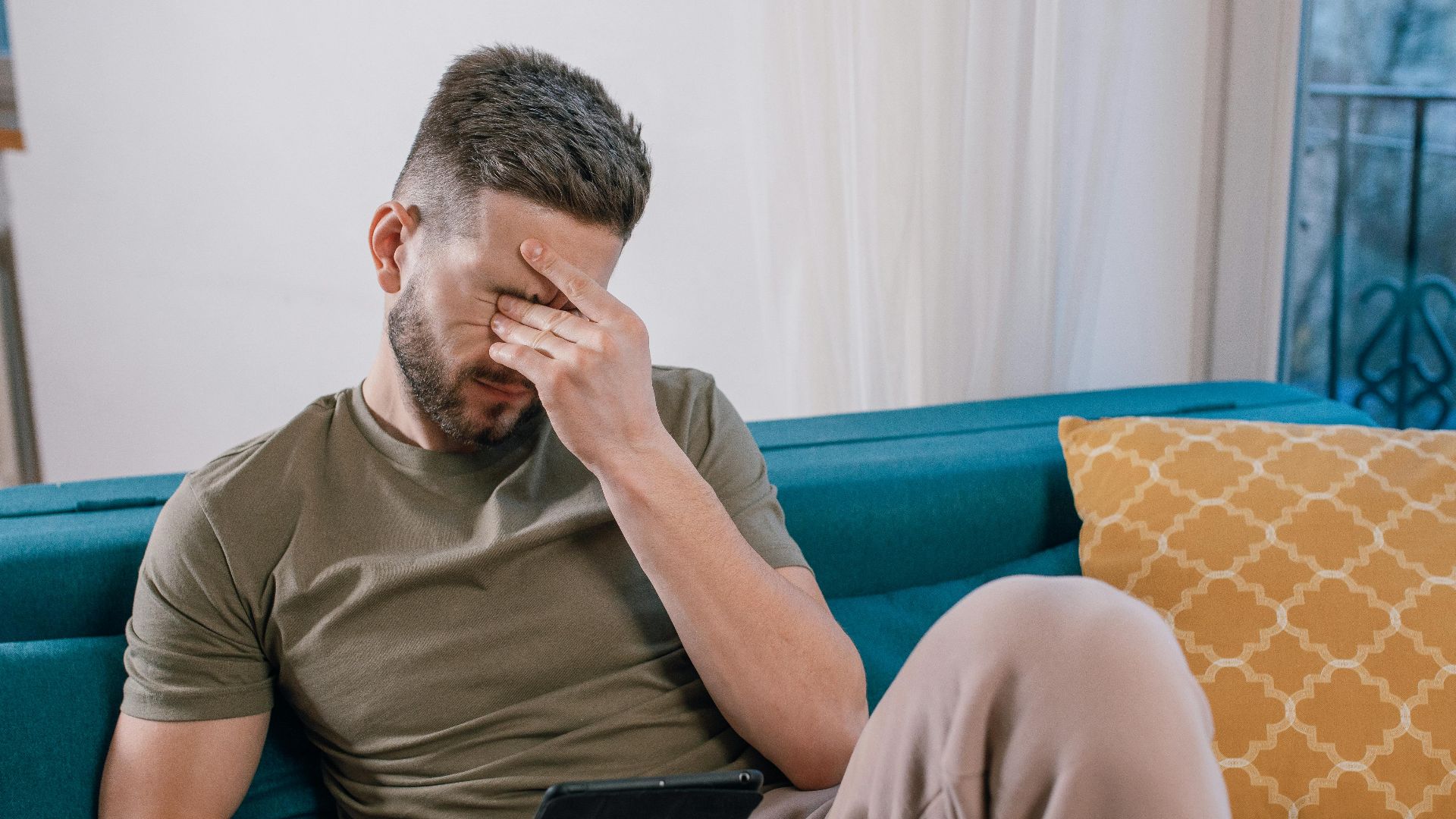 A man sits on a sofa holding a tablet, visibly stressed and covering his face with one hand.