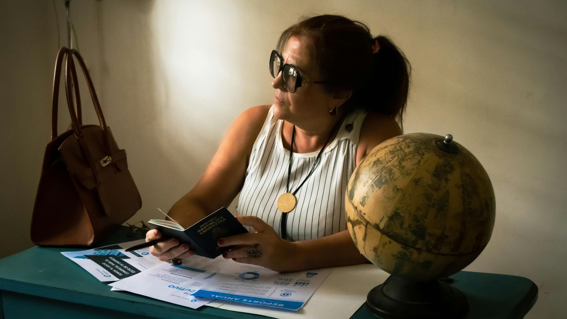 A woman seated indoors reviews travel documents beside a globe, suggesting travel planning.