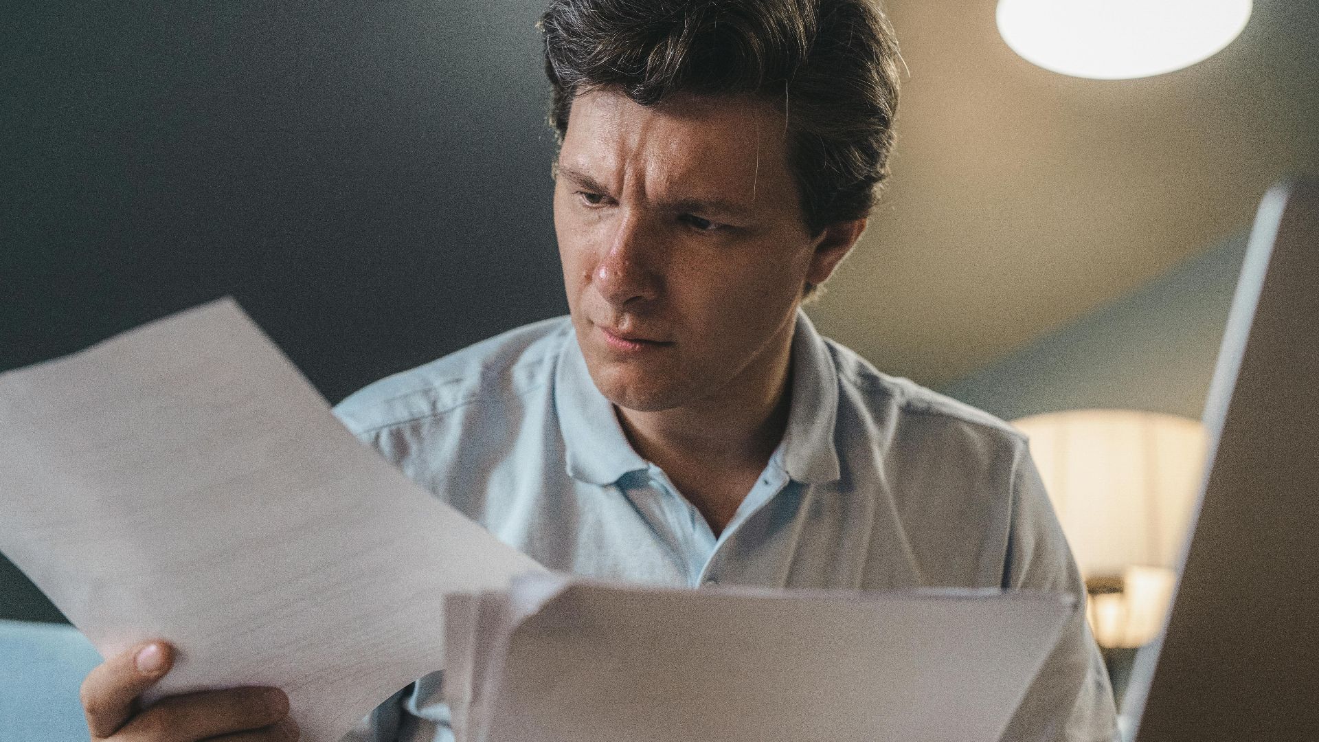 A man intently examines papers, seated indoors under warm lighting, focusing on his work.