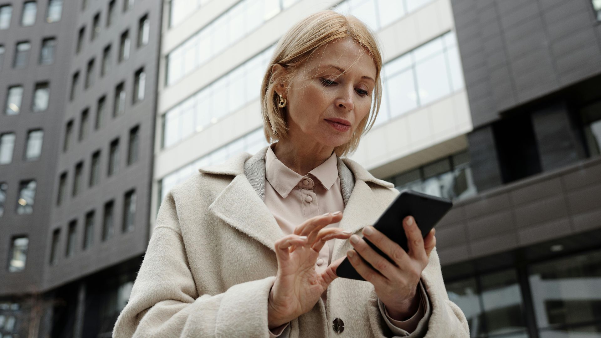 Woman in coat using smartphone in front of modern building. Professional and focused expression.