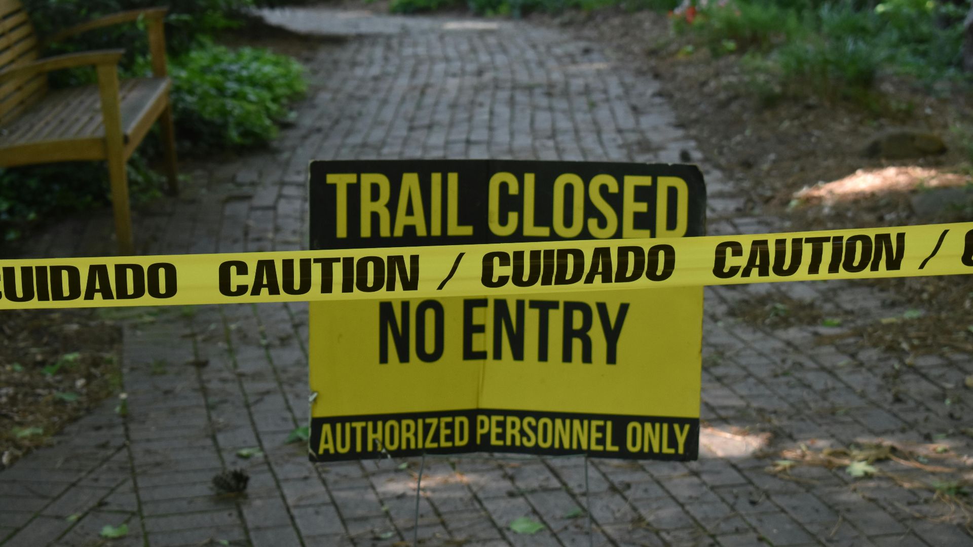Trail closed sign with caution tape blocking pathway in a park setting.
