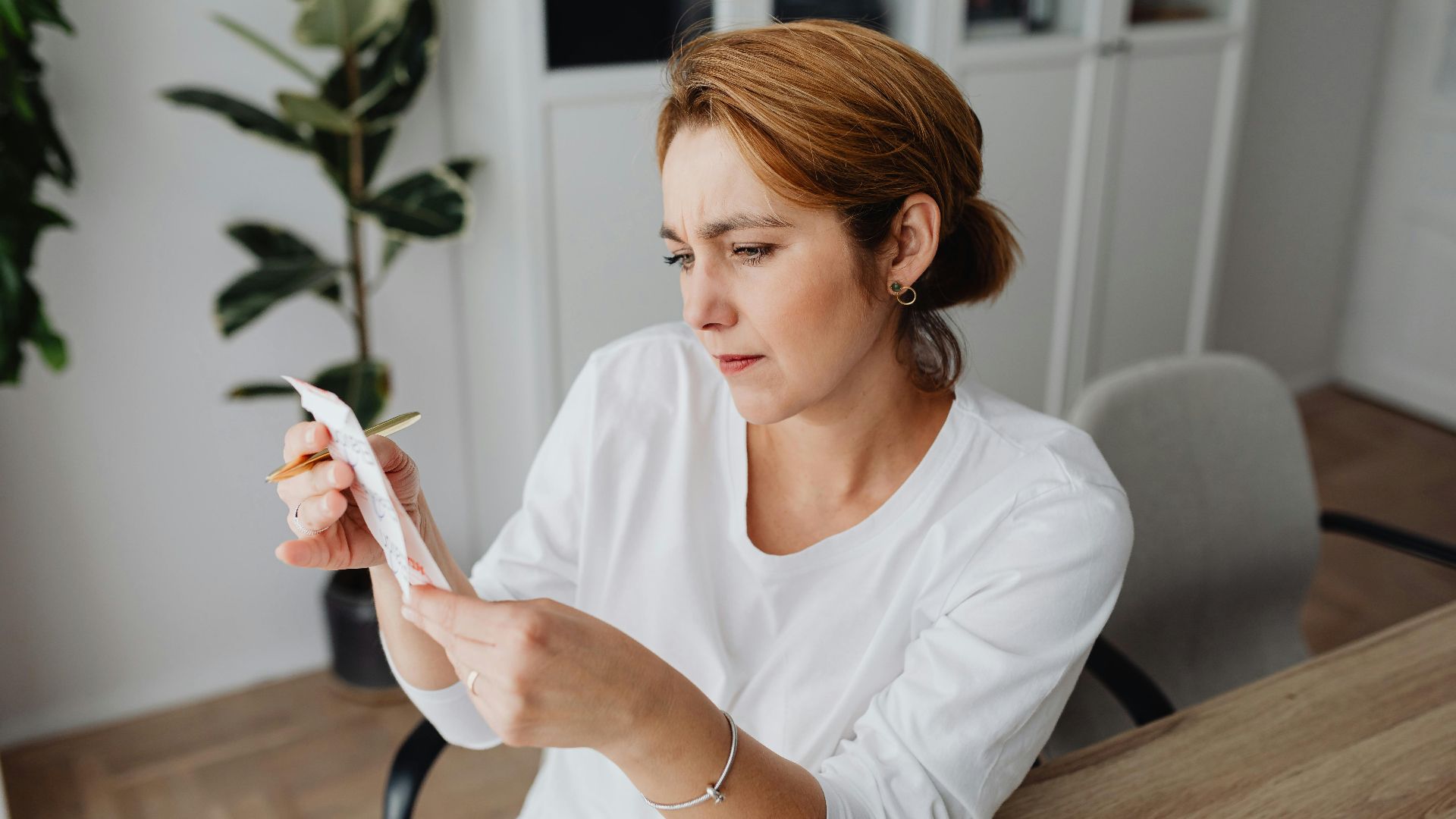 A concerned adult woman examines a receipt while sitting at her office desk, indicating financial scrutiny.