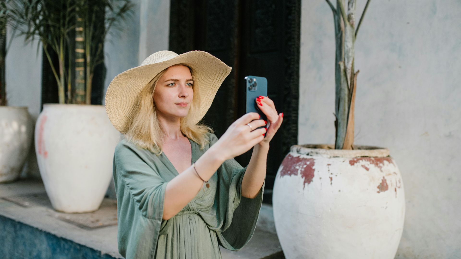 Female tourist wearing summer dress and broad brim hat taking photo on cellphone white standing on street near palms growing in clay jugs