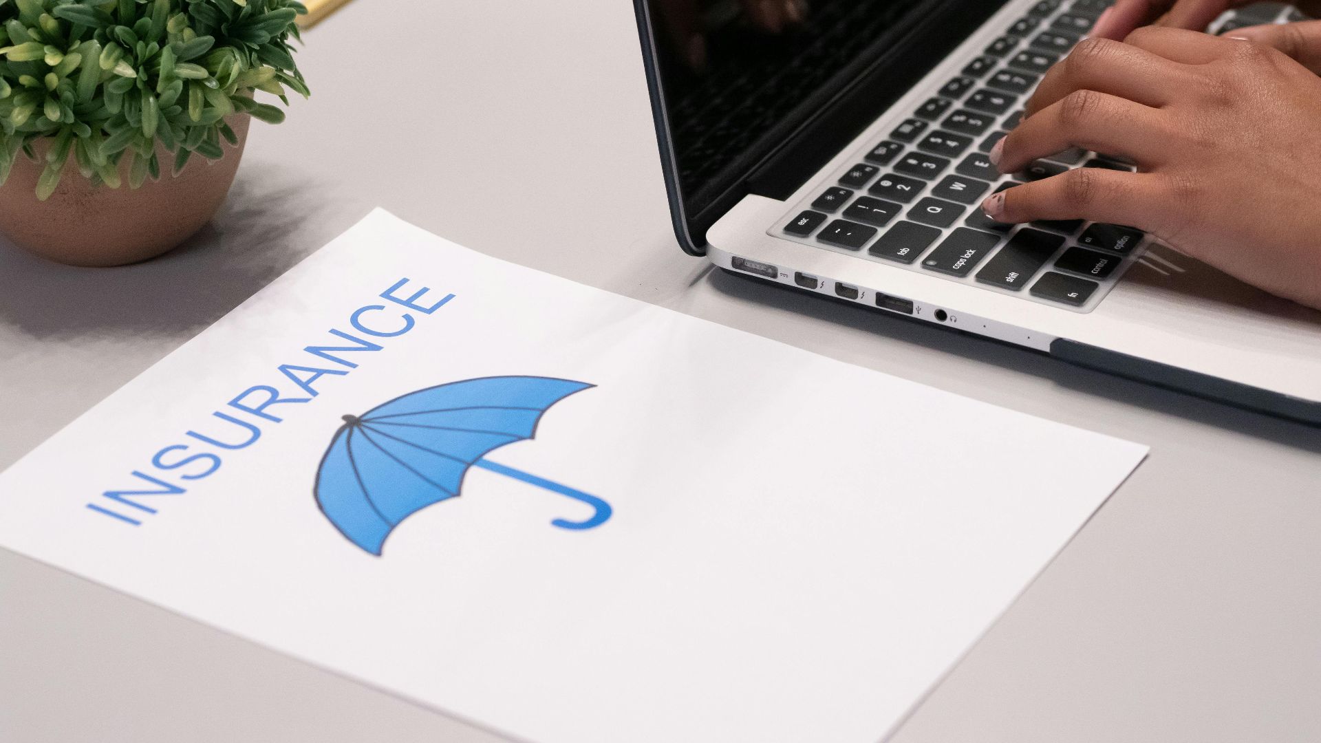 Close-up of hands typing on laptop with an insurance document visible on the desk.