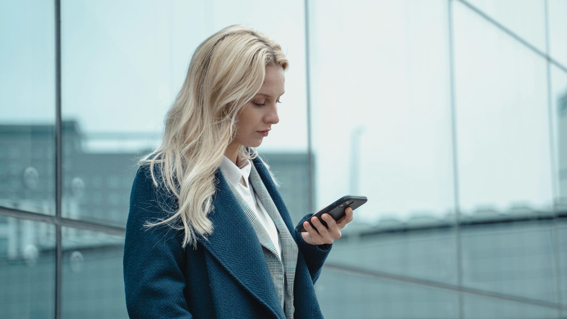 Professional woman in blue coat using smartphone outside office building.