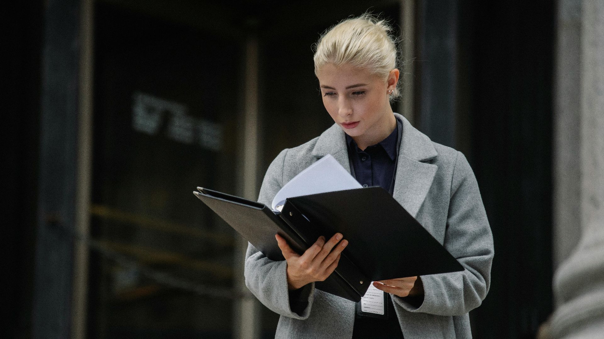 Focused young businesswoman in formal clothes standing outside modern building and reading notes in folder