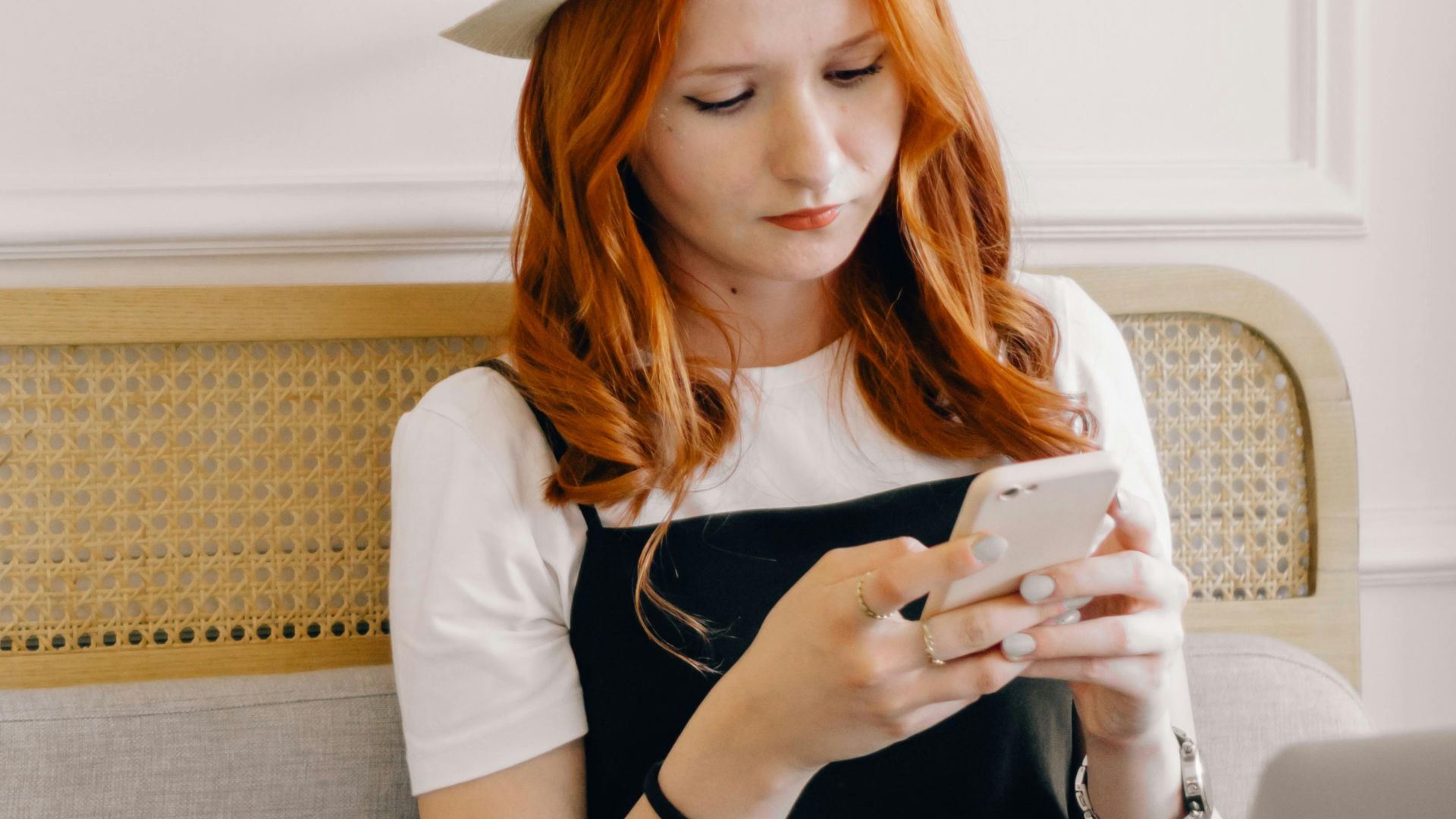 A young woman in a fedora hat texting on her smartphone while sitting with a laptop indoors.