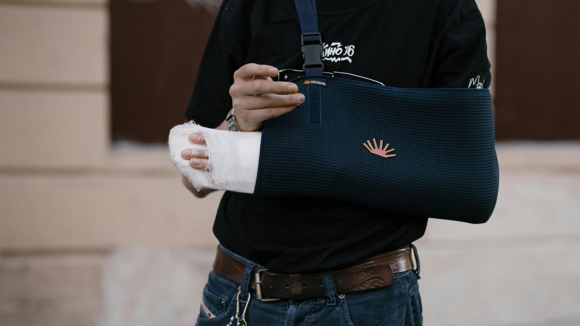 A man wearing a dark sling with a bandaged arm, standing outdoors in casual attire.