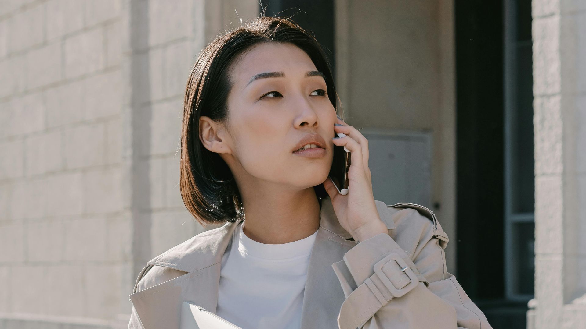Confident Asian businesswoman in trench coat making a phone call while walking outside an urban building.