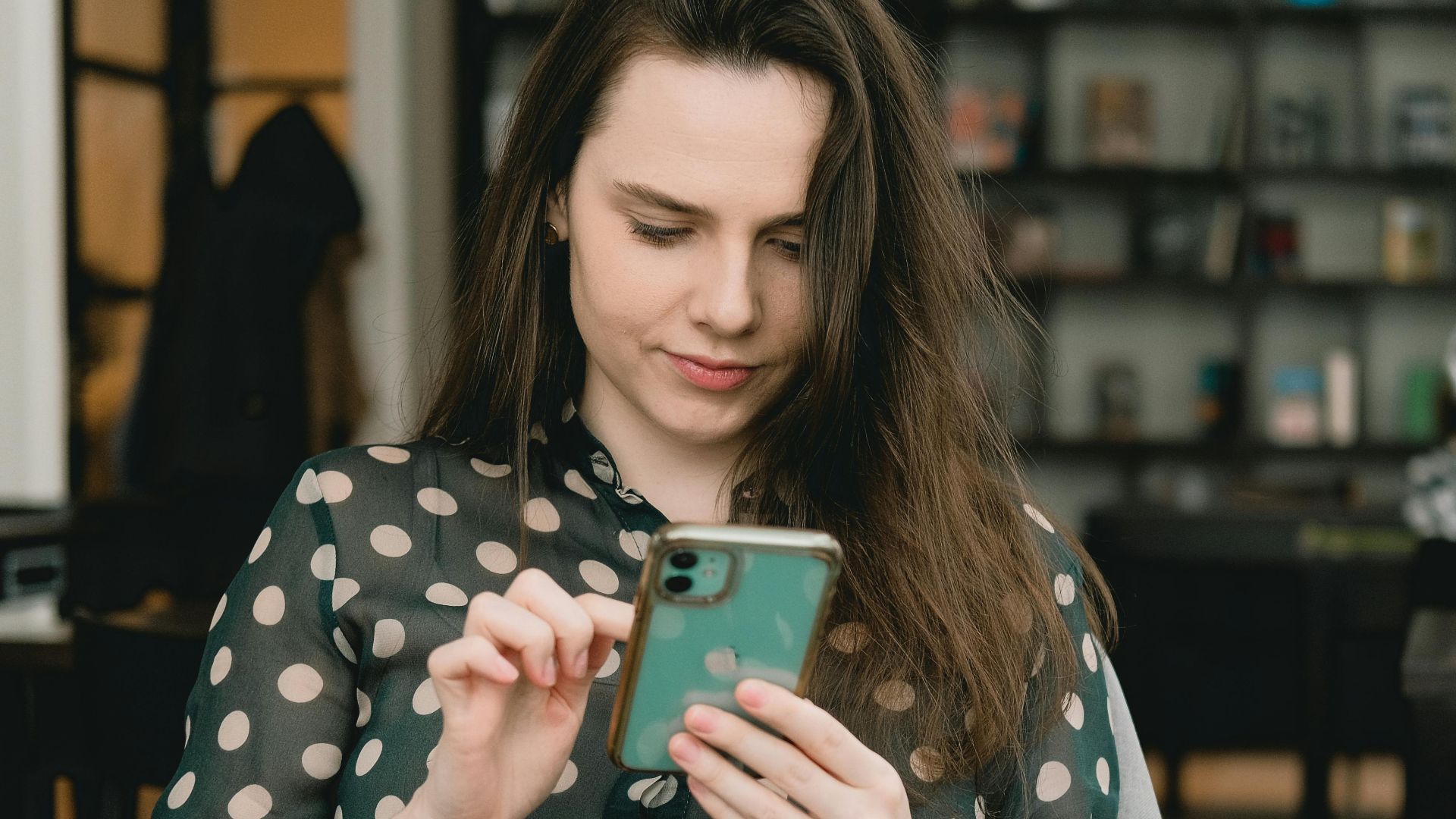 Woman in polka dot dress using smartphone indoors at a library. Cozy and modern setting.