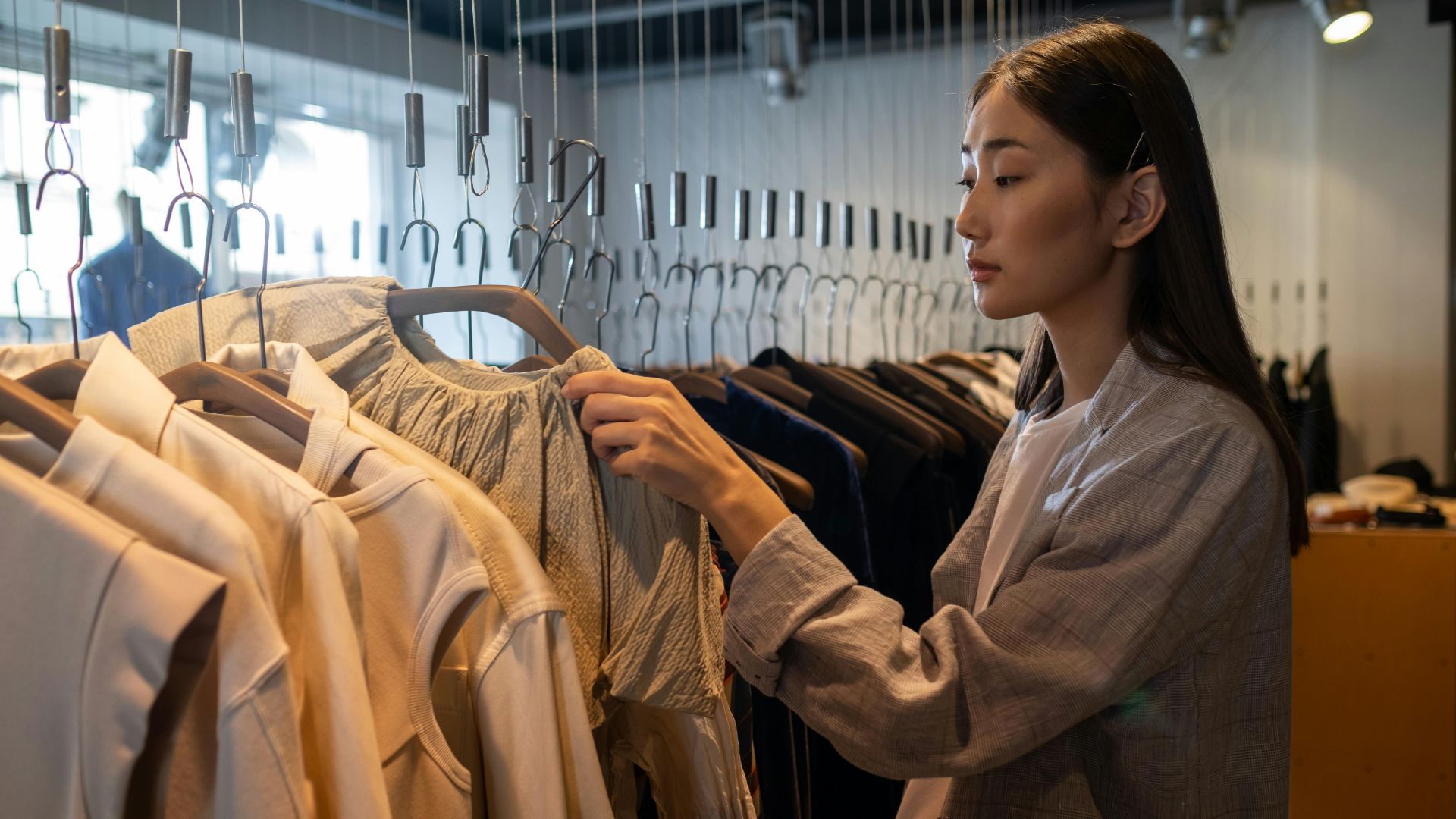An Asian woman browsing clothing in a boutique shop, examining fashion items on hangers.