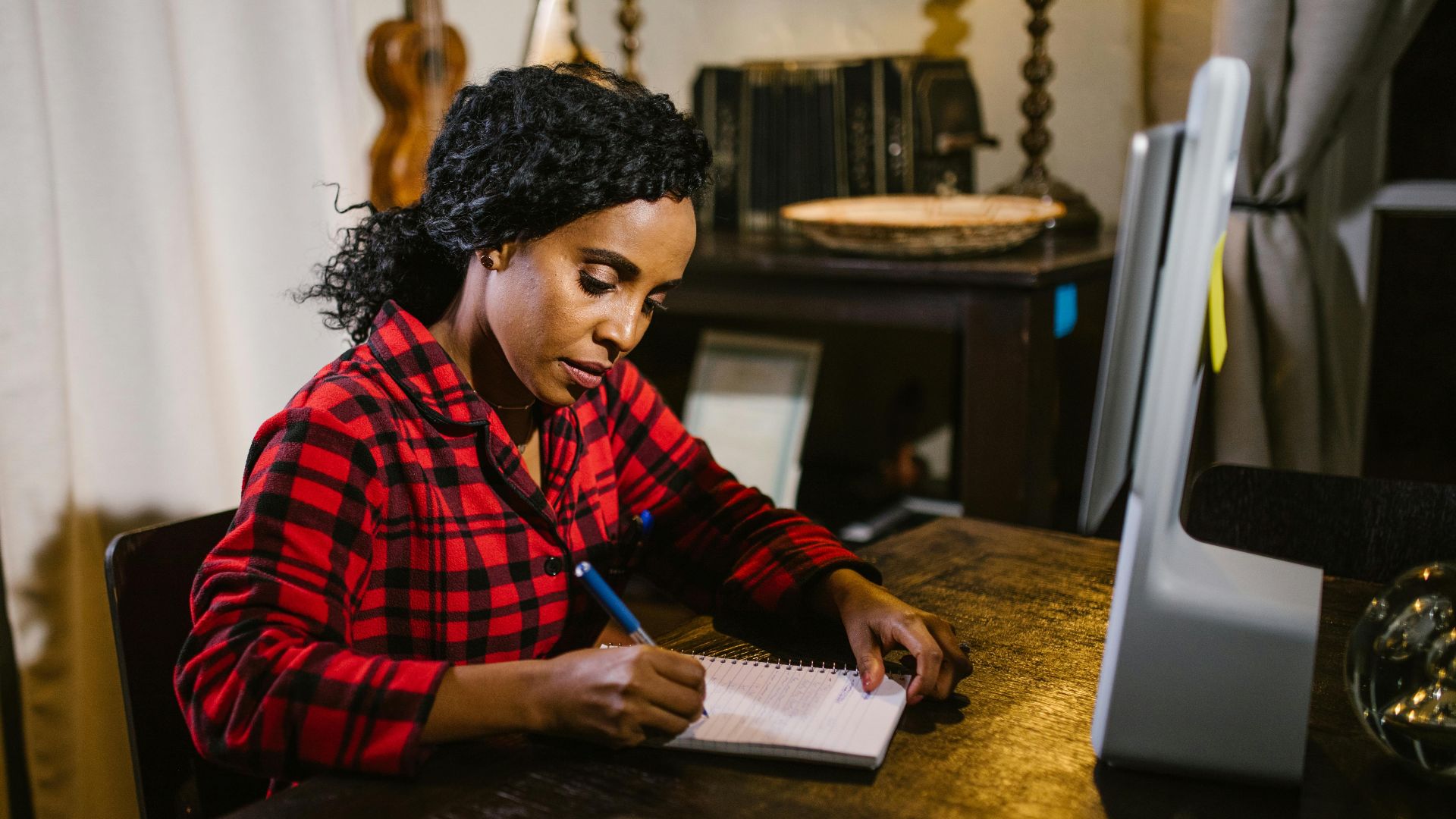An African American woman focuses on writing notes at her home workspace.