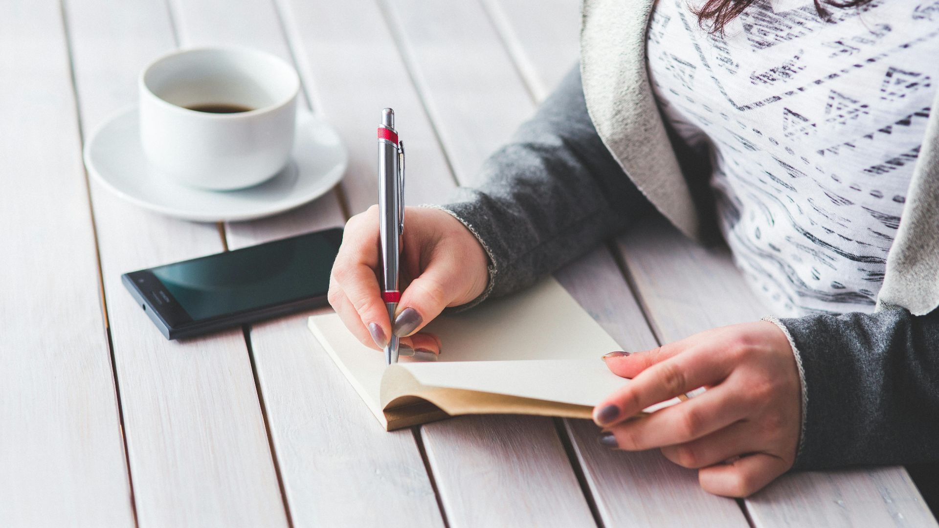 A woman writes in a journal next to a cup of coffee and a smartphone on a wooden table.